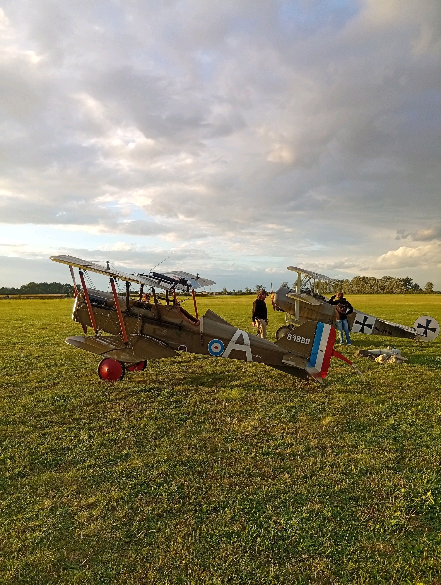 In Szewce, Poland, two biplanes are parked on a grassy field under a cloudy sky. The first biplane, olive green with red wheels, is in the foreground and appears to be a vintage model. A roundel insignia is visible on its fuselage. Behind it, another biplane, also olive green, is partially visible. Two individuals stand near the second biplane; one wearing a dark hoodie and jeans, the other in a light-colored shirt and trousers. The environment is a rural setting with a treeline in the background. The lighting suggests it is either early morning or late afternoon.