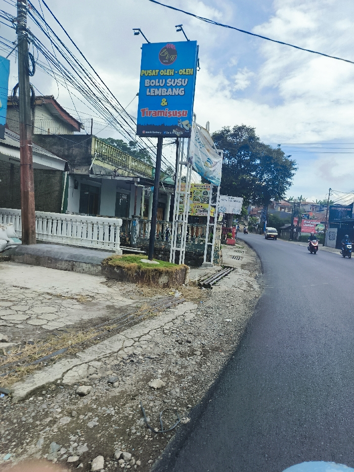 The image shows a street in Lembang, Indonesia. It's a narrow, paved road with a small, cracked concrete shoulder on the right side. A large sign advertising "Pusat Oleh-Oleh Bolu Susu Lembang & Tiramisu" is positioned at the edge of the shoulder. There are several houses and buildings visible on the left side of the road, with a white picket fence separating the road from the buildings. A few trees are visible in the background, along with a few other signs and a car parked in a driveway. Two motorbikes are visible in the distance on the right side of the road.