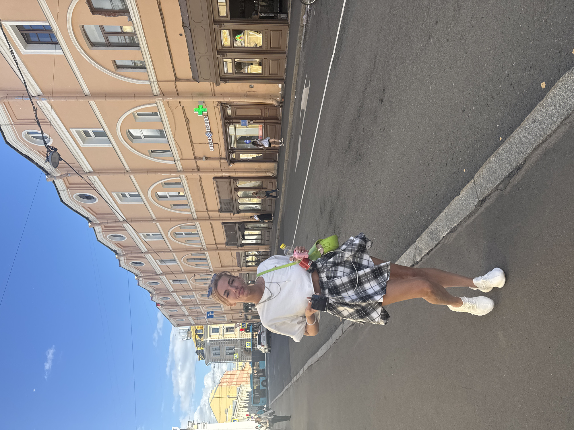 A young Caucasian woman stands on a sidewalk in front of a peach-colored building with arched windows, likely in Saint Petersburg, Russia, given the Russian text on the building signage. She is wearing a white crop top, a black and white plaid skirt, and white sneakers. She holds a phone in her left hand and a green purse with a water bottle and other items in her right hand. The street in front of the building appears to be a one-way street with traffic and a bus in the background. The sky is blue with some clouds.