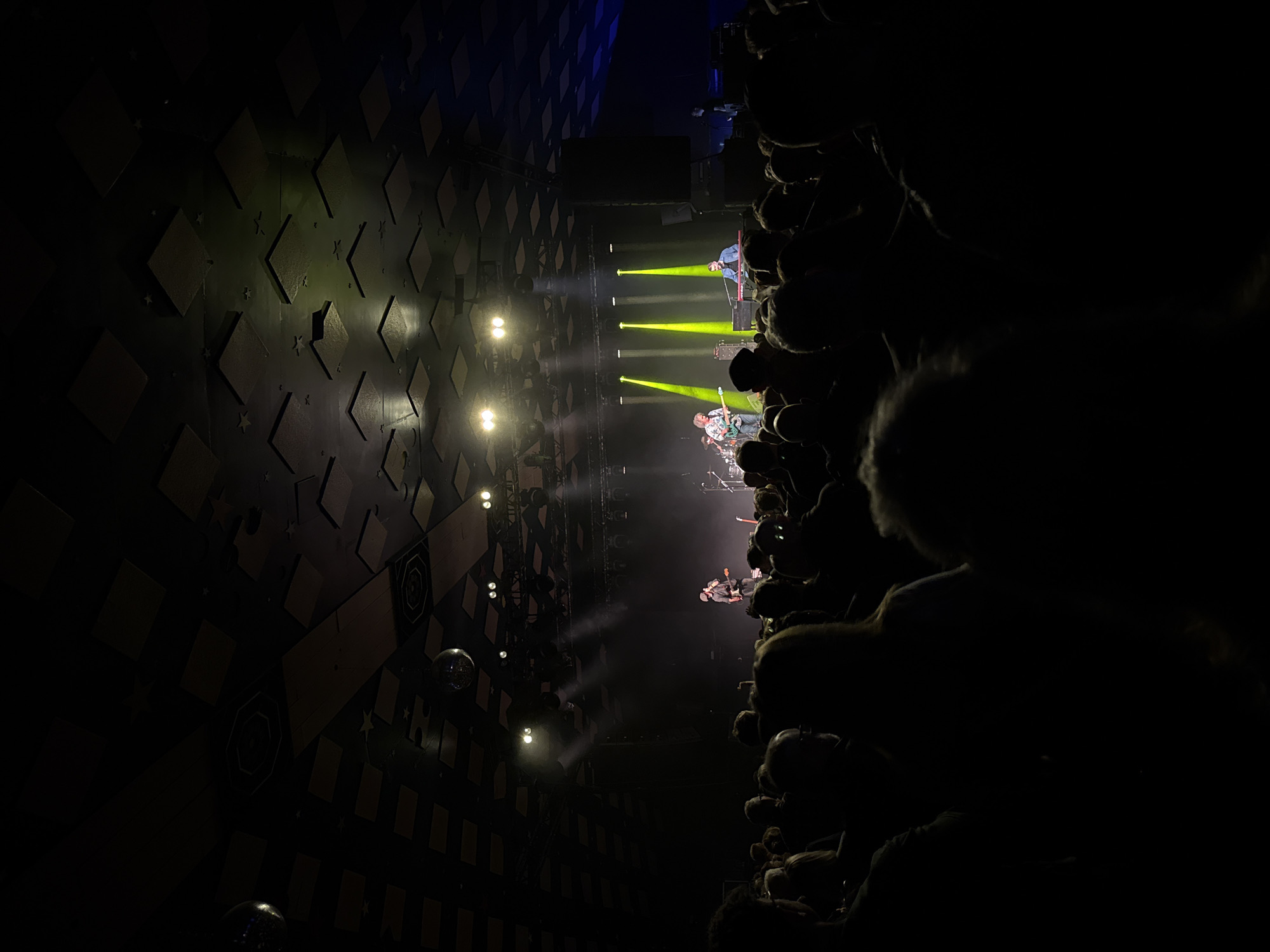 The image captures an indoor live music concert from an audience perspective. The specific location within the city of Unknown, Unknown is not discernible. In the foreground and midground, a dense crowd of individuals is silhouetted, facing a brightly lit stage in the background.
On the stage, four musicians are visible. From left to right: a man in a cap and jacket plays an electric guitar; another man in a lighter shirt also plays an electric guitar; a third man in a blue shirt is seated at a red keyboard/synthesizer; and a fourth figure is partially obscured on the far right. The stage is illuminated by several prominent vertical yellow spotlights and overhead white stage lights. Large, rectangular black speaker cabinets are visible on the right side of the stage.
The venue's ceiling is dark, featuring a textured design composed of square and rectangular acoustic panels. Two reflective disco balls are suspended from the ceiling in the upper left, and small star-shaped decorative elements are scattered across the dark ceiling surface. Some blue accent lighting is visible on the far-right ceiling panels.
