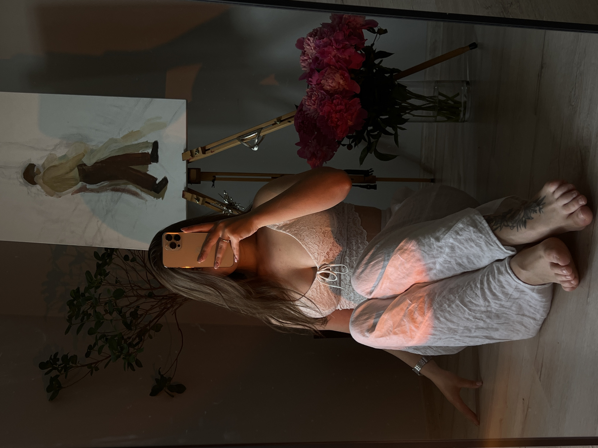 A young woman, partially visible in a mirror's reflection, sits on a wooden floor. She is wearing a white lace bralette and loose white pants, with a watch on her left wrist. Her right leg is bent, and her left leg is extended. She holds a gold smartphone in her right hand, obscuring her face. Beside her, on the floor, is a vase filled with vibrant pink peonies. Behind her, an easel holds a partially finished painting of two figures embracing. A potted plant with green leaves stands to the left of the easel. The scene is illuminated by warm, diffused light casting long shadows. The location is not specified within the image.