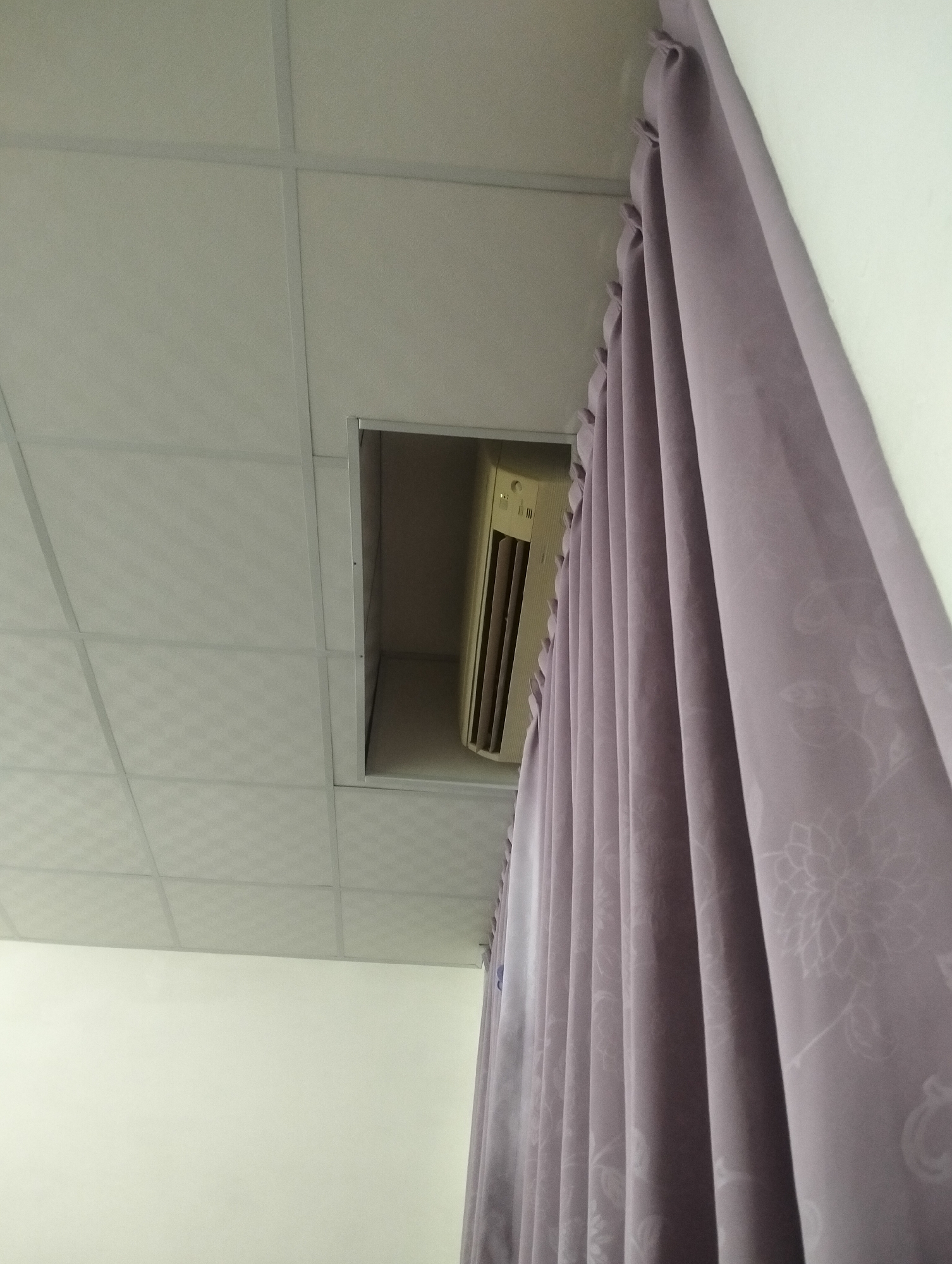 An indoor scene shows a drop ceiling with a rectangular opening revealing an air conditioning unit. To the right, a portion of a light purple curtain with a floral pattern is visible, hanging against a white wall. The overall impression is of a functional, possibly institutional or residential space. The image's content does not provide information specific to Kaohsiung, Taiwan, or any particular city.