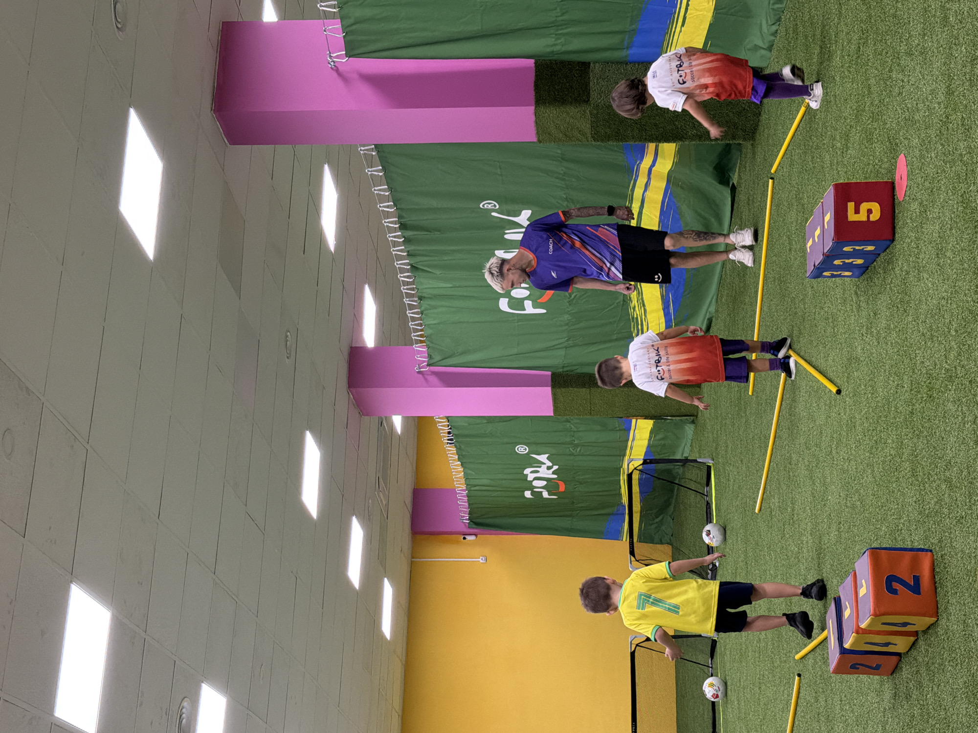 A group of people are in an indoor sports facility. There are several children and at least one adult, likely a coach. The children are wearing athletic clothing, including soccer jerseys and shorts, and appear to be participating in a soccer training session. The adult is wearing a purple and orange jersey with "COACH" printed on it.
The scene is set on an artificial turf field, indicated by the green carpet. There are various training props visible, including yellow poles, small soccer goals, and colorful foam cubes stacked to form numbered blocks. One cube has a "5" on it, another has "3", and a third has "21". A pink disc is placed near the "5" cube.
The facility has a bright and well-lit interior, with white ceiling lights visible. The background features green and yellow fabric banners, one of which has a logo that reads "FORK" with "soccer & life skills" underneath. Another banner has colorful stripes. The walls are painted in muted tones of pink and yellow.
The activity appears to be a structured training or practice session, possibly for young soccer players. The participants seem engaged, with one child in a yellow jersey with the number "7" on it actively participating. The lighting and indoor setting suggest the activity is taking place during the day, regardless of external weather conditions. There are no explicit emotional cues visible, but the organized setup implies a focused and possibly instructive environment.