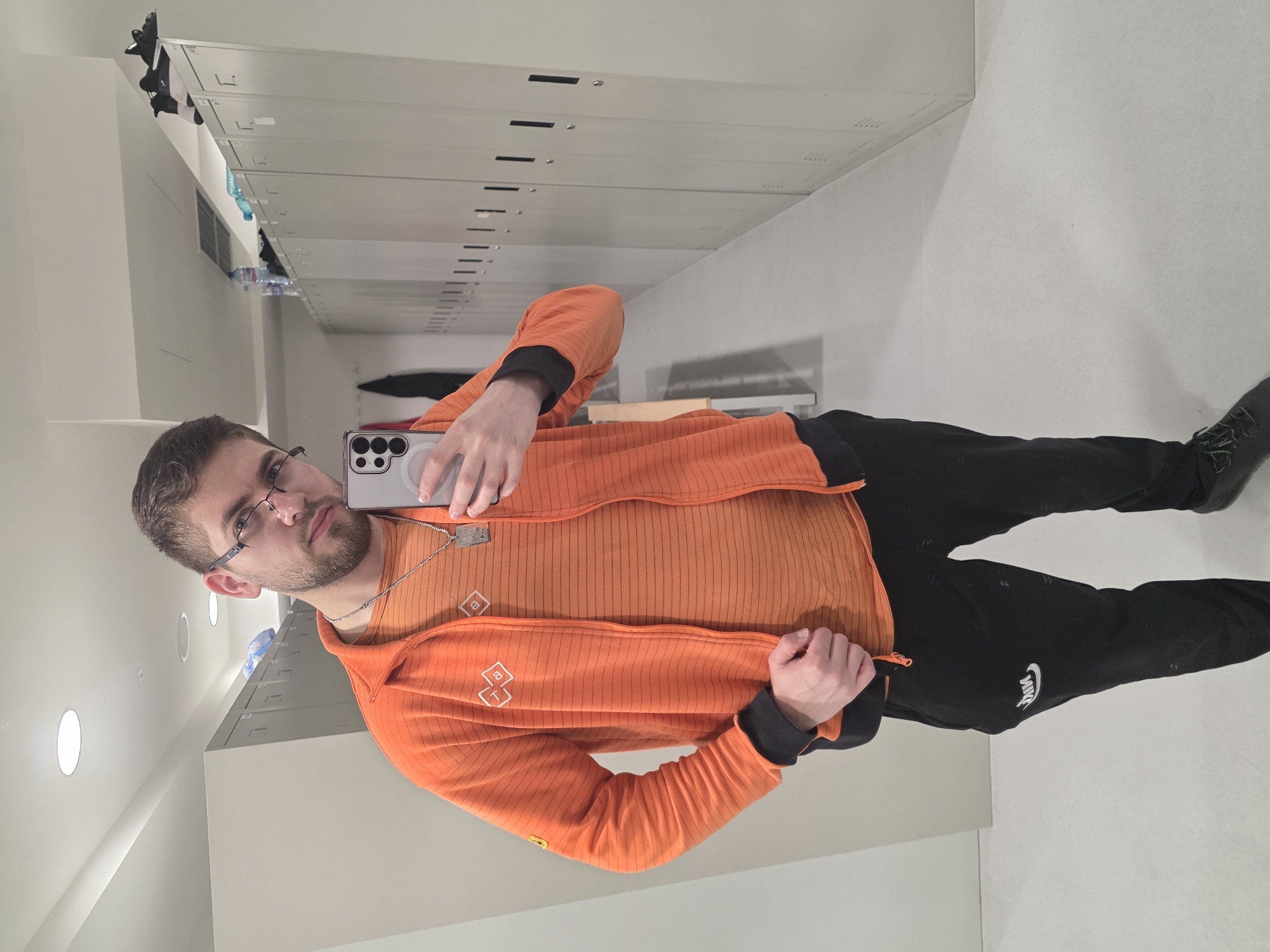 The image is a selfie of a man in a locker room or hallway with lockers on one side. He is wearing glasses, an orange zip-up jacket, and black sweatpants with a Nike logo. He is holding a smartphone, presumably taking the picture. The environment appears to be a sterile, institutional setting with rows of lockers. There is no evidence to suggest it is Plovdiv, Bulgaria.