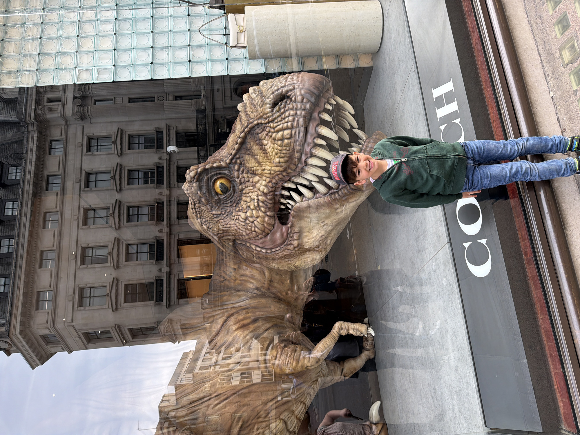 A young boy stands next to a large, realistic Tyrannosaurus Rex model displayed behind a shop window. The boy wears a green jacket, blue jeans, and a baseball cap. He is smiling and looking toward the camera. The T-Rex model is detailed with sharp teeth, textured skin, and visible claws. The shop window behind the model displays the word "COACH" vertically. The reflection in the window shows a building exterior with windows and architectural details, as well as a cloud-filled sky. The location is the city of London, United Kingdom.