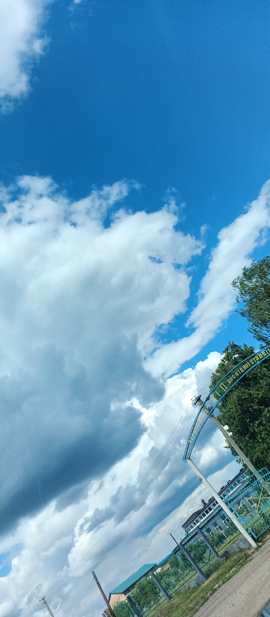 The image is a low-angle, wide shot of a sky filled with dramatic clouds, ranging from bright white cumulus to darker, more ominous storm clouds. A bright blue sky is visible in the upper portion of the frame. In the foreground, a blue arched gateway with yellow lettering is visible, partially obscured by lush green trees. The text on the gateway appears to be in Ukrainian and reads "МТП ПО ДСОПІДНОЇ СТАНЦІЇ," which translates to "MTF PO DSOPIDNE STATION." To the right of the gateway, a multi-story building with many windows can be seen, suggesting an institutional or research facility. A green metal fence runs along the property line in front of the building. Electrical poles and wires are also present in the scene, indicating utility infrastructure. The ground in the foreground is a dirt or gravel surface. The overall scene suggests an outdoor environment, possibly on the grounds of an agricultural or research station in the city of Kholodnianske, Ukraine, under a dynamic and changing sky. No people are visible in the image.