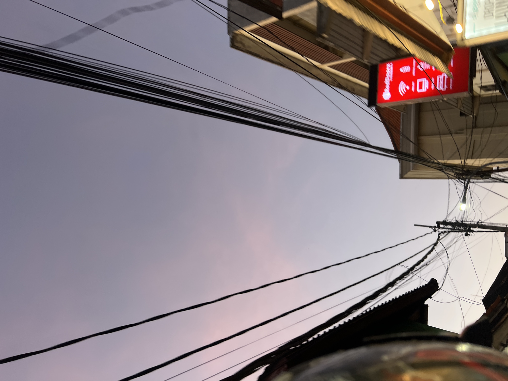 An outdoor, low-angle, medium shot captures a residential street in Kuta, Indonesia, during twilight. The sky transitions from a pale pink near the horizon to a soft lavender overhead. A dense network of black power lines crisscrosses the frame, casting faint shadows and creating a dynamic visual texture against the subdued sky.
On the right side of the image, a building with a light-colored facade and reddish-brown horizontal slatted accents is visible. Attached to the building is a brightly lit red sign displaying the "RedDoorZ" logo in white. Below the logo are white icons representing a Wi-Fi signal, a smartphone, a laptop, and a printer. To the right of this sign, another illuminated sign is partially visible, displaying white text on a light background.
On the left side of the image, the edge of a dark, peaked roofline is visible, suggesting a dwelling. In the lower left corner, a reflective, metallic surface, possibly part of a helmet or vehicle, occupies a portion of the frame, tinted with hues of yellow and orange from ambient light. A streetlamp with a visible bulb is illuminated on a utility pole in the mid-ground, casting a soft light. The overall scene evokes a quiet, urban evening in a residential area.
