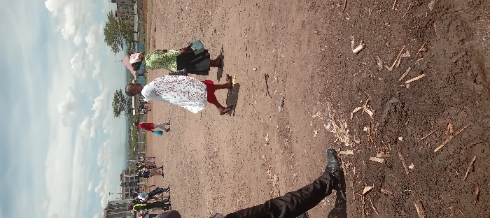 The image shows a dirt field with scattered sticks and debris. The field is located in the city of Kitengela, Kenya, at the coordinates -1.47 latitude and 36.959 longitude. There are a number of people walking across the field, including two women wearing brightly colored clothing. In the background, there is a building, trees, and a cloudy sky.