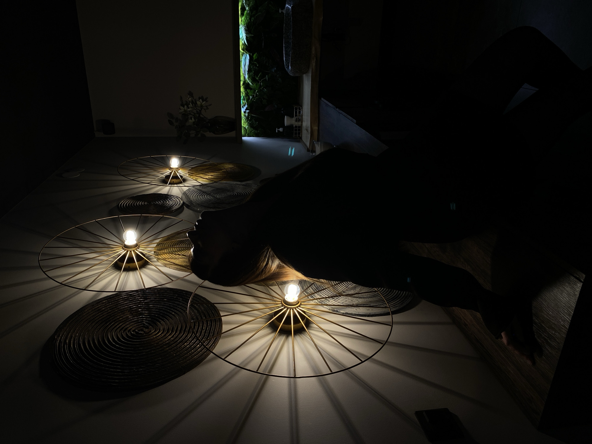 A dimly lit room features several circular, spoked light fixtures on the ceiling, illuminating sections of the floor. Dark, circular objects, seemingly decorative mats, are positioned around the light fixtures. The silhouette of a person appears to be lying on the floor, partially obscuring the light fixtures. A doorway in the background reveals a space with green foliage. The location of the scene in the city of Votkinsk, Russia is indeterminable from the image.