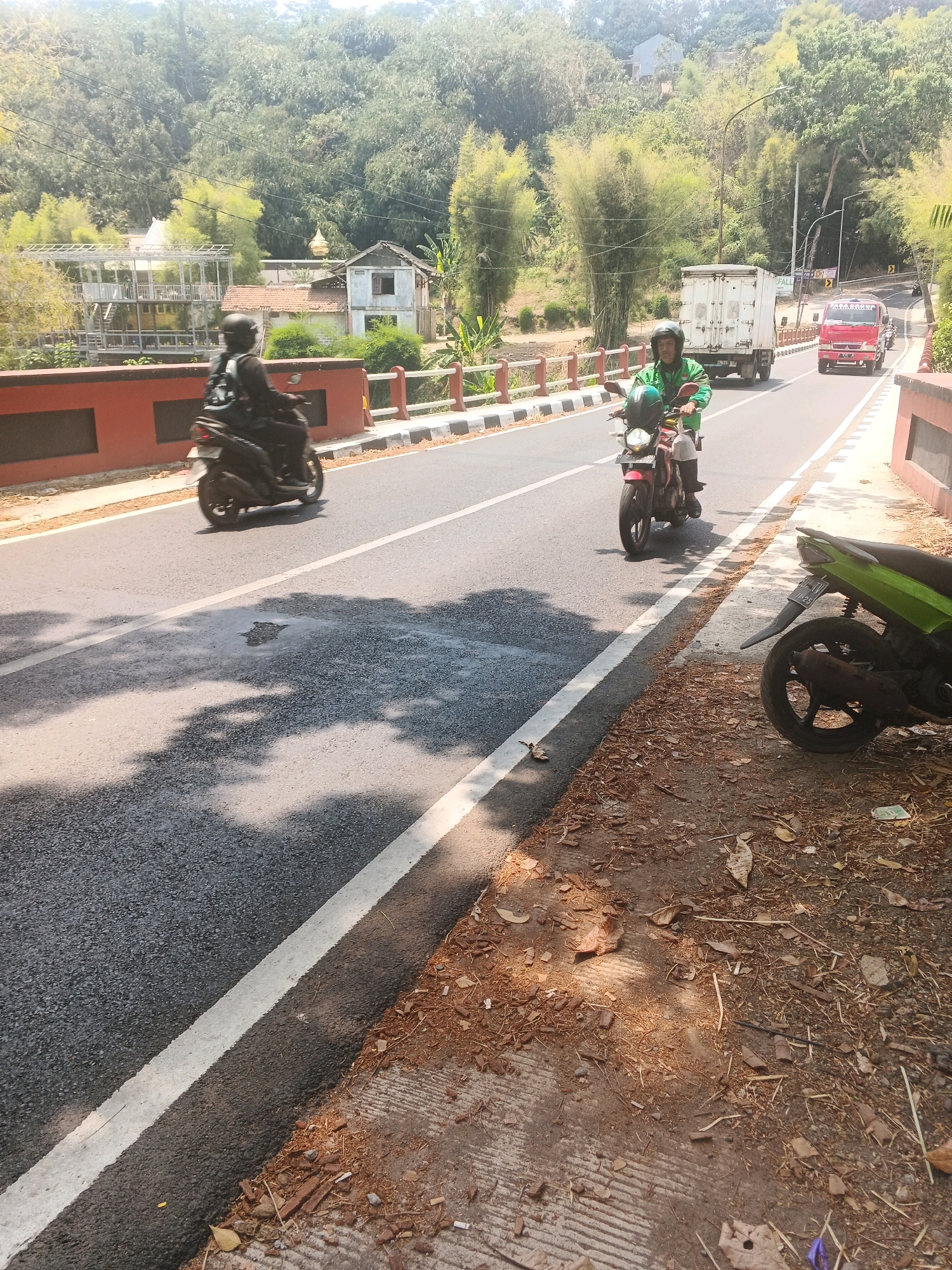 In Semarang, Indonesia, a street scene unfolds with two motorcycles and two trucks on a paved road bordered by a red barrier on the left and a grassy embankment with scattered dry leaves and debris on the right. A person on a black motorcycle, wearing a helmet and backpack, rides in the left lane. Further ahead in the right lane, another person on a red motorcycle, also wearing a helmet and a green jacket, is in motion. Behind them, a white truck and a red truck are visible on the road. The background features a lush, green hillside densely covered with trees and bamboo, with a few buildings nestled among them. The sunlight casts shadows across the road and embankment.