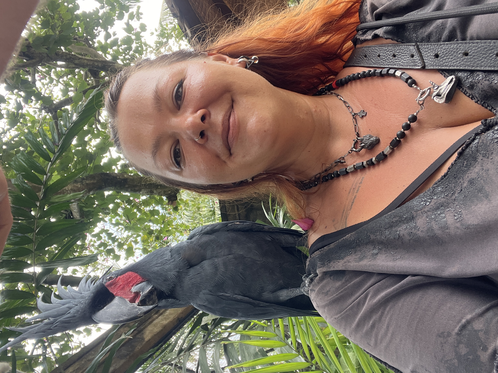 A woman with reddish-orange hair, fair skin, and visible piercings is holding a large black palm cockatoo. The woman is wearing dark-colored clothing and necklaces. The cockatoo has a large beak with red facial skin. The background consists of lush green tropical foliage, indicating a tropical or forest-like setting. The image was likely taken in the city of Sukawati, Indonesia.