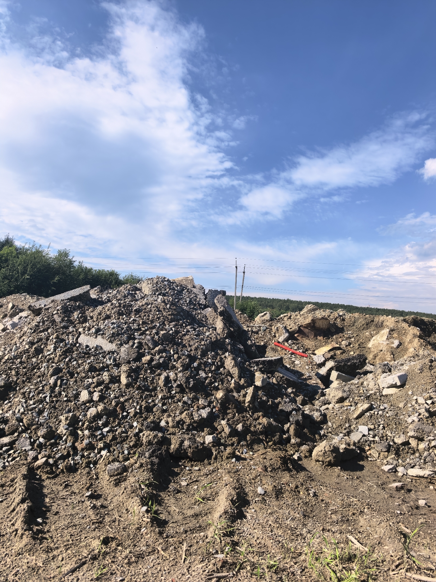 The image depicts a construction debris pile under a bright blue sky with scattered white clouds. The pile is composed of concrete, asphalt, rocks, and dirt, with some larger pieces of broken concrete clearly visible. In the background, a dense green forest borders the debris. Several power lines stretch horizontally across the frame, supported by utility poles on the left. A red pole or stick is embedded in the debris on the right side of the image. The ground in the foreground shows tire tracks, indicating recent activity. The scene is brightly lit, suggesting it is daytime. The weather appears clear and sunny. No people are visible in the image. There is no visible text.