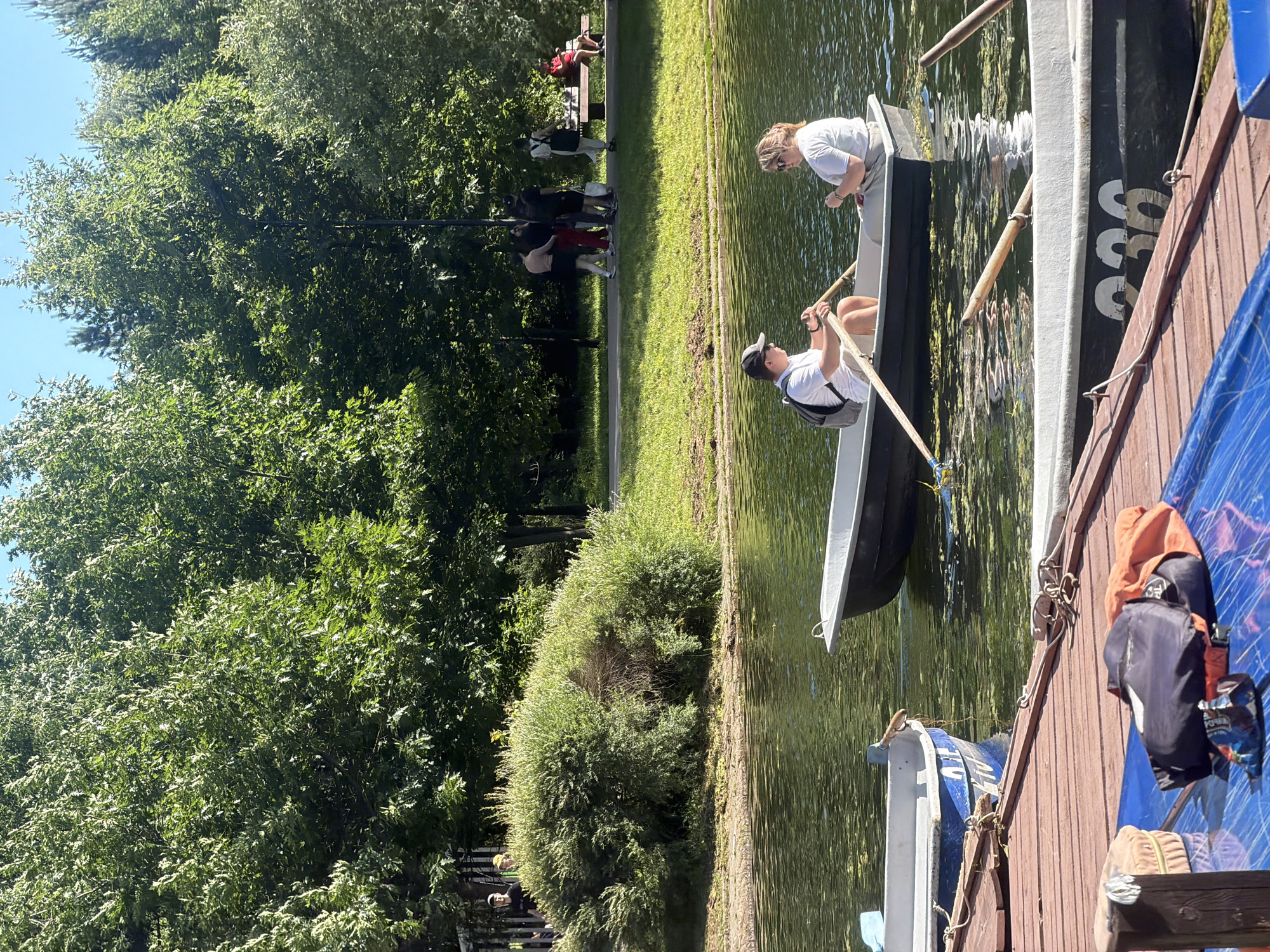 Two people are rowing a boat on a lake. The person in the front, wearing a white t-shirt and a black baseball cap, is holding an oar. The person in the back, wearing a white t-shirt and sunglasses, is also holding an oar. They are both looking forward. The boat is white and black. In the background, there are trees and a path with several people walking. One person is sitting on a bench. The lake is green and there is some algae on the surface. There are other boats docked on the left side of the image. The scene appears to be a park on a sunny day. The location is the Friendship of Peoples Park in Khimki, Russia.