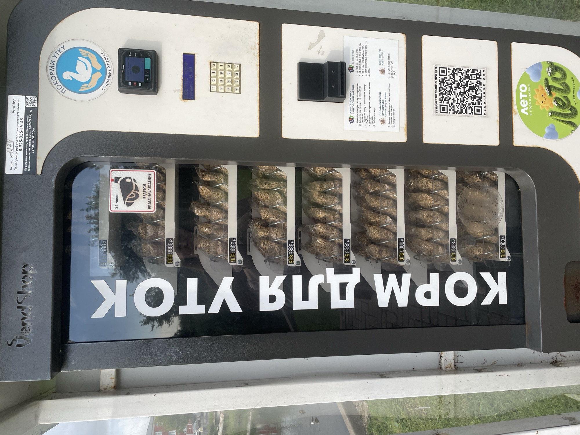 This is a photograph of a vending machine for duck food, likely located in the city of Sergiyev Posad, Russia, as indicated by the presence of Russian text on the machine and a QR code for VEND-SHOP.COM. The machine is vertical, with a glass display case showing rows of bagged duck food. Large white lettering on the glass reads "КОРМ ДЛЯ УТОК" (KORM DLYA UTOK), meaning "FEED FOR DUCKS". Each product slot displays a price, such as "9:10 8888P" and "9:15 8888P", indicating the currency is likely Russian Rubles (P).
To the right of the food display are operational instructions and payment options. There is a keypad with numbers and letters, a card reader, and a slot for cash or coins. A sign with a swan and hands, featuring the text "ПОКОРМИ УТКУ" (POKORMI UTKU - "FEED THE DUCK") and "СОЦИАЛЬНЫЙ ПРОЕКТ" (SOTSIAL'NYY PROEKT - "SOCIAL PROJECT"), is prominently displayed. Below this, there is a digital display and a camera icon labeled "24 ЧАСА" (24 CHASA - "24 HOURS") and "ВЕДЕТСЯ ВИДЕОНАБЛЮДЕНИЕ" (VEDETSYA VIDEONABLYUDENIYE - "VIDEO SURVEILLANCE IS BEING CONDUCTED").
A QR code for VEND-SHOP.COM is present, along with a section displaying information in Chinese, suggesting the machine might also cater to Chinese tourists. Another prominent graphic at the bottom depicts a sunny, cartoon-like landscape with the text "ЛЕТО В ПОДМОСКОВЬЕ" (LETO V PODMOSKOV'YE - "SUMMER IN THE MOSCOW REGION"), reinforcing the likely Russian location. The background shows a blurry glimpse of greenery and what appears to be a body of water with ducks, consistent with a park or public space where ducks are found. The machine itself is gray and appears to be mounted on a white structure.