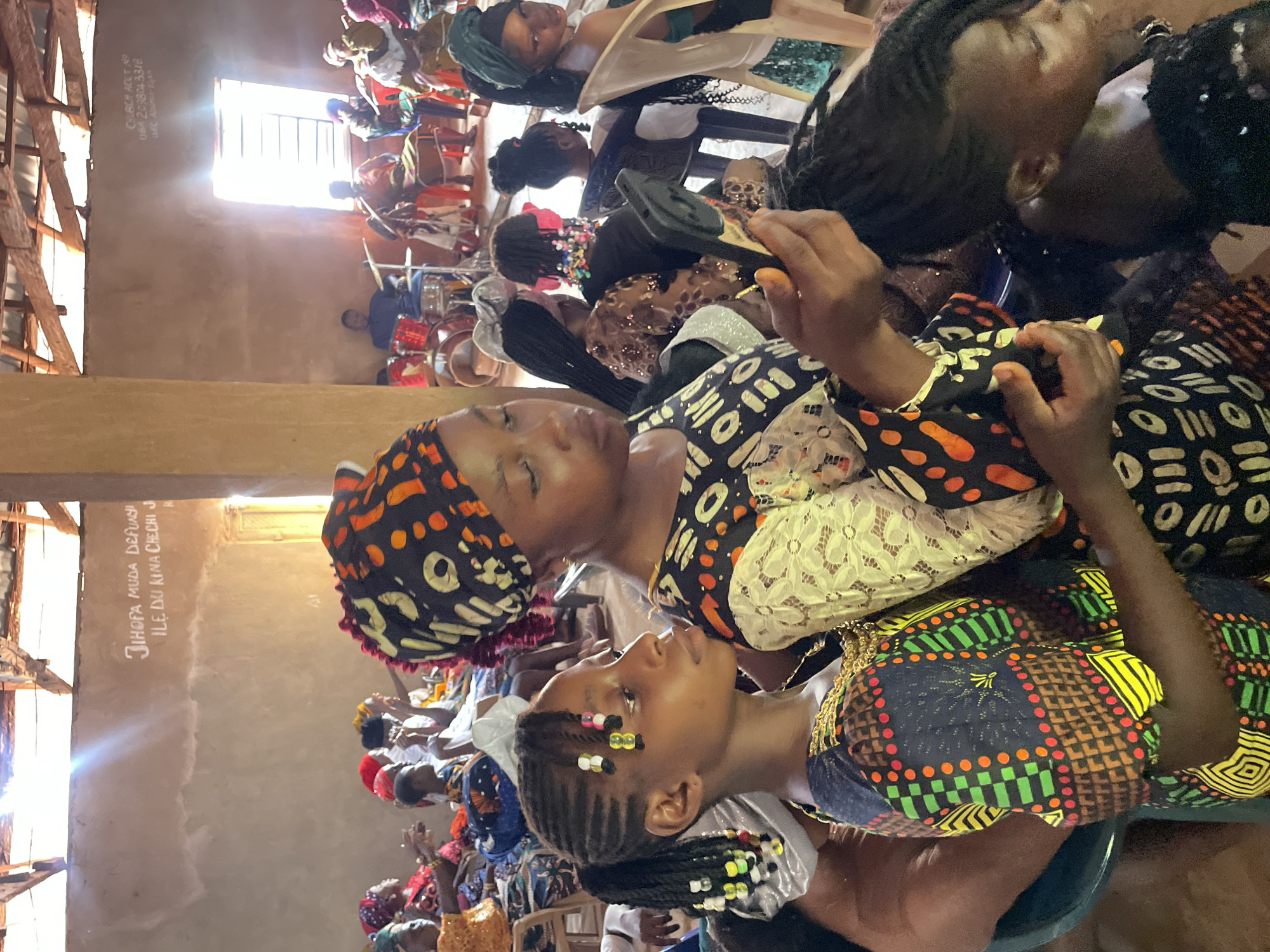 The image captures an indoor scene of a religious gathering in Ogbe, Nigeria, with a group of people, mostly women and children, attending a church service. Individuals are dressed in vibrant African prints and headscarves. Some are seated and appear to be listening or participating in the service. A group of drummers are visible in the background, and writing on the wall says "JIHOFA MUDA DEFUWI ILE DU KIMA CHECHI JI". Natural light filters through a window. The overall ambiance suggests a cultural event or religious ceremony within a modest building.