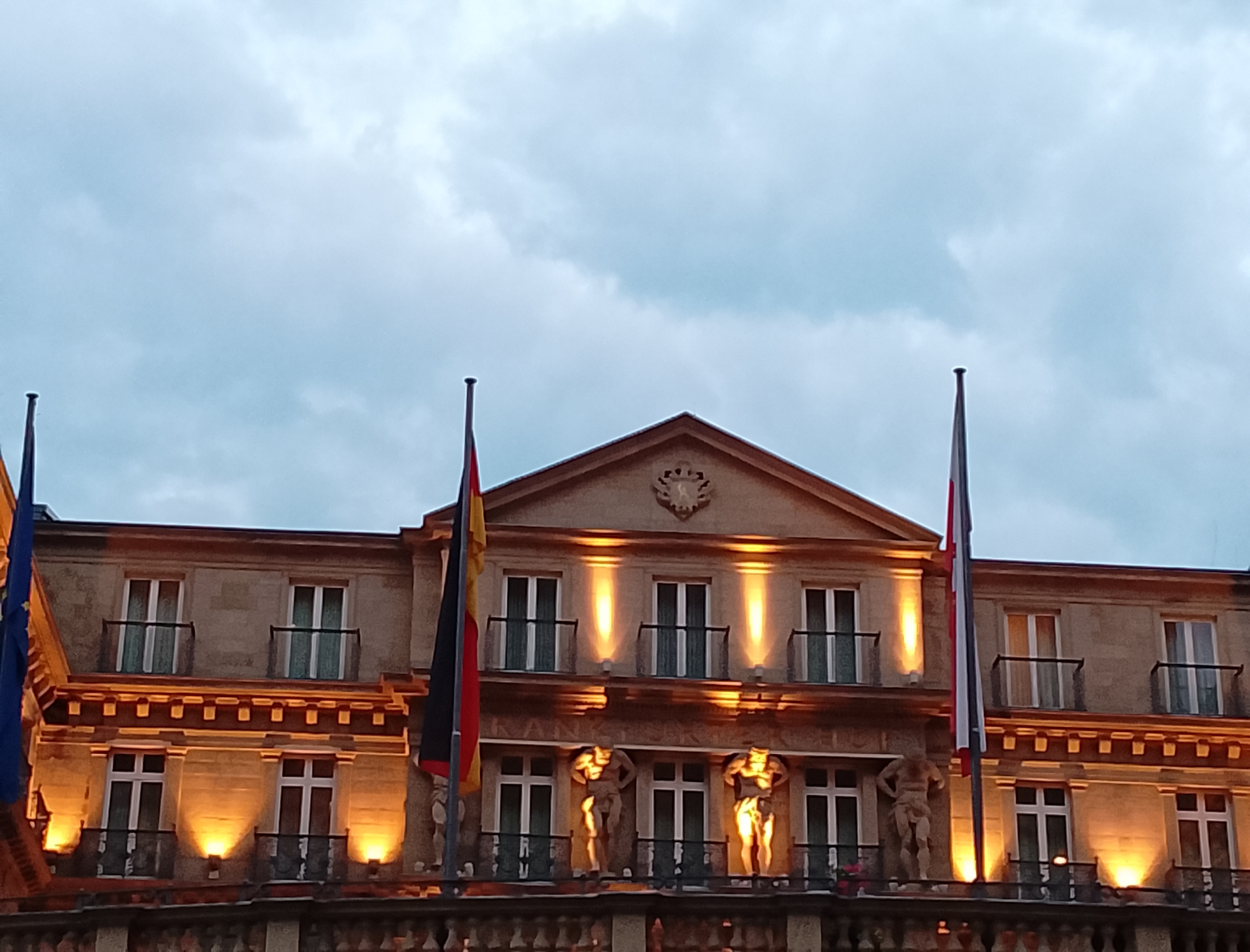 The image shows the facade of a multi-story building. The building has a classical architectural style, with ornate detailing and numerous windows. On the facade, there are two statues of male figures holding up balconies. The building is illuminated by warm, artificial lighting. Two flags are visible on flagpoles, one on the left and one on the right side of the building. The flag on the left appears to be a blue and yellow European Union flag, while the flag on the right is a tricolor flag with red, white, and blue vertical stripes. The sky above is overcast with clouds. The overall scene suggests a formal or governmental building, possibly at dusk or night.
