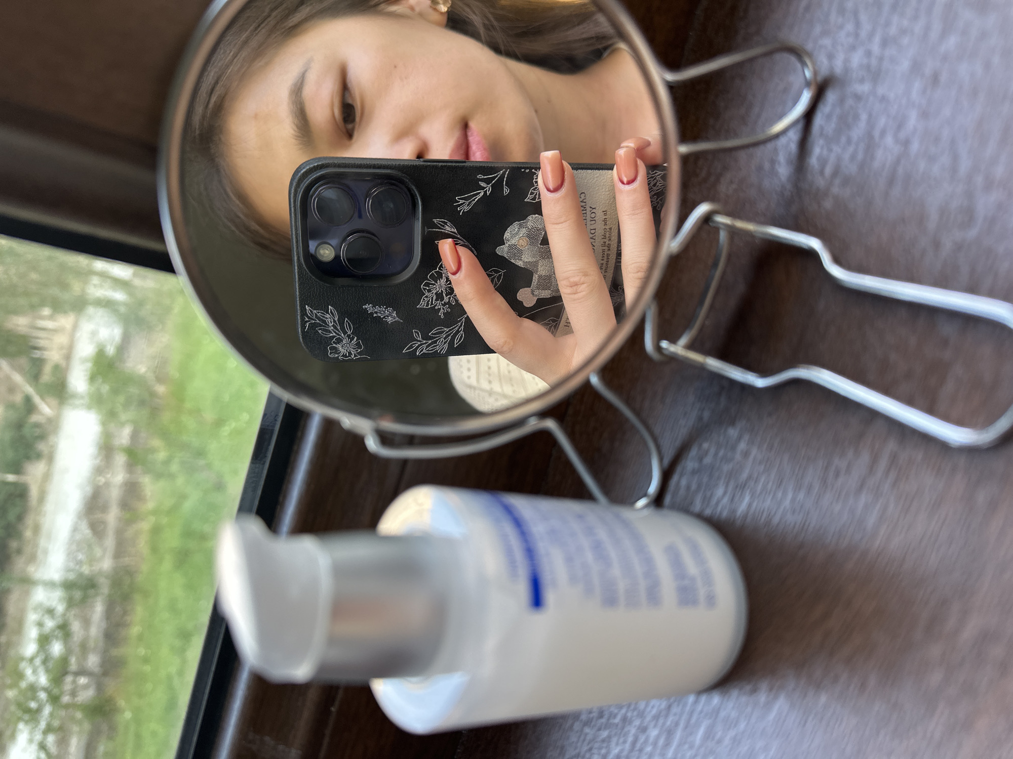 A young woman is taking a selfie in a handheld mirror. She is looking at her phone, which she is holding up to take the picture. The phone has a black case with a floral design. Her nails are painted a light brown color with dark red tips. She is wearing a light-colored sweater. Next to her is a white bottle with a pump dispenser. The background is blurred, showing greenery and what appears to be a road or path. The scene is set on a wooden surface. There is no information available to determine the location in the city of Кызыл-Булак, Kyrgyzstan.