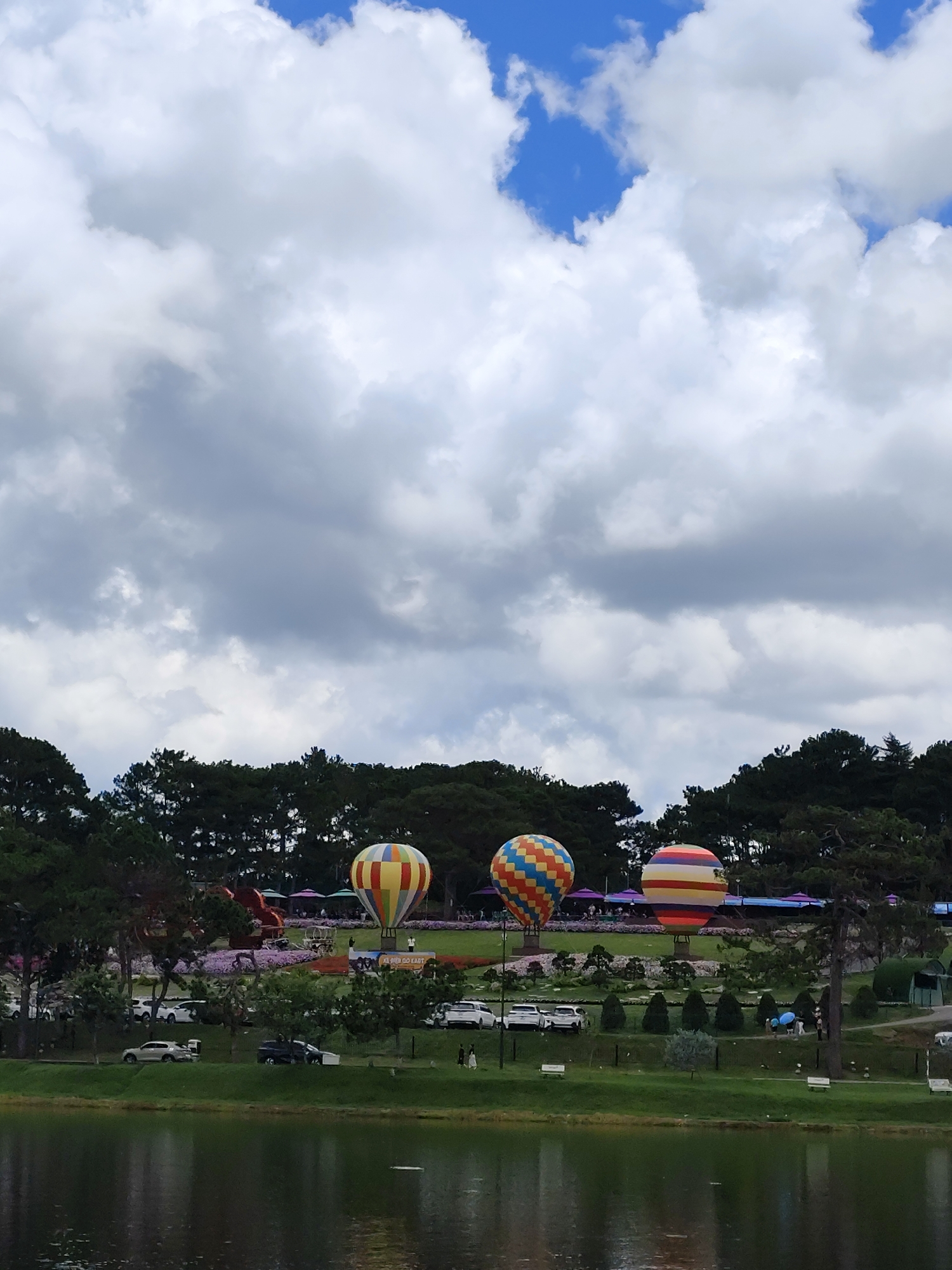 The image depicts a scenic view of Xuan Huong Lake in Da Lat, Vietnam, on a cloudy day. In the foreground, a calm lake reflects the sky and surrounding trees. Beyond the lake, a manicured park area is visible with lush green grass, scattered trees, and well-maintained gardens. Several hot air balloons, featuring colorful geometric patterns, are prominently displayed in the park, suggesting a festival or event. The balloons are anchored to the ground and are spaced apart. In the background, a dense line of pine trees borders the park, characteristic of Da Lat's landscape. A few vehicles are parked along a road within the park. Several people are scattered throughout the park, some walking and others observing the scenery or the hot air balloons. The overall atmosphere is serene and picturesque.
