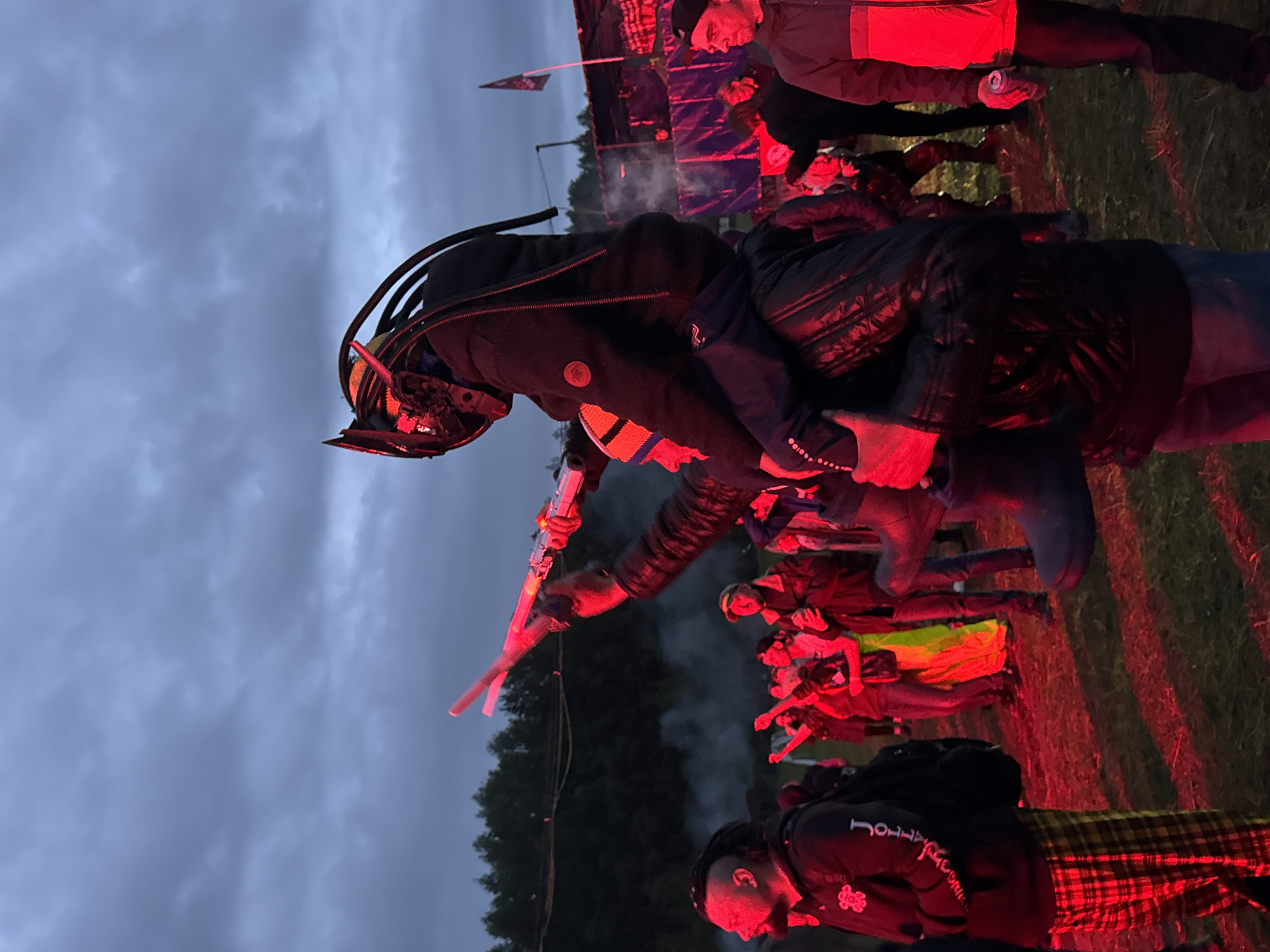 The image depicts an outdoor gathering taking place in Bolshaya Kivara, Russia, under an overcast, twilight sky. The scene is heavily illuminated by artificial red light, casting a dominant crimson hue across the foreground and midground. The ground consists of uneven dirt and sparse patches of grass.
In the center of the frame, a person wearing a dark, ribbed jacket and a distinctive helmet resembling a "Predator" alien, complete with dark dreadlock-like appendages, is perched on the shoulders of another individual. This lower individual wears a dark, possibly padded or leather jacket and a striped beanie (orange, blue, and white). Their right arm is extended upwards, holding two glowing, cylindrical objects that emit red/pink light, resembling light sticks or toy sabers. Their left arm is wrapped around the legs or waist of the person on their shoulders, seemingly for support.
To the left of the central figures, a man with dark skin and dreadlocks styled in an updo wears a dark hoodie with white lettering (partially visible as "JO...") and red and yellow plaid pants. He carries a dark backpack on his left shoulder and is looking towards the right.
To the far right, a man wearing a dark baseball cap and a two-toned jacket (dark on top, red on bottom) holds a silver beverage can in his left hand, resting at his side.
In the midground, several other individuals are visible, also bathed in the red light. Some are seen with their arms raised, possibly dancing or reacting to an event. One person in the midground is notably wearing a bright, possibly neon green or yellow garment. Wisps of white smoke or haze are present in this area, catching the red light.
In the background, a dense line of dark evergreen trees extends across the horizon. To the far right, a dark structure, possibly a stage or temporary shelter, is discernible, with a dark flag (featuring a lighter emblem) flying from a pole near it. The overall atmosphere suggests an outdoor concert, festival, or rave event.