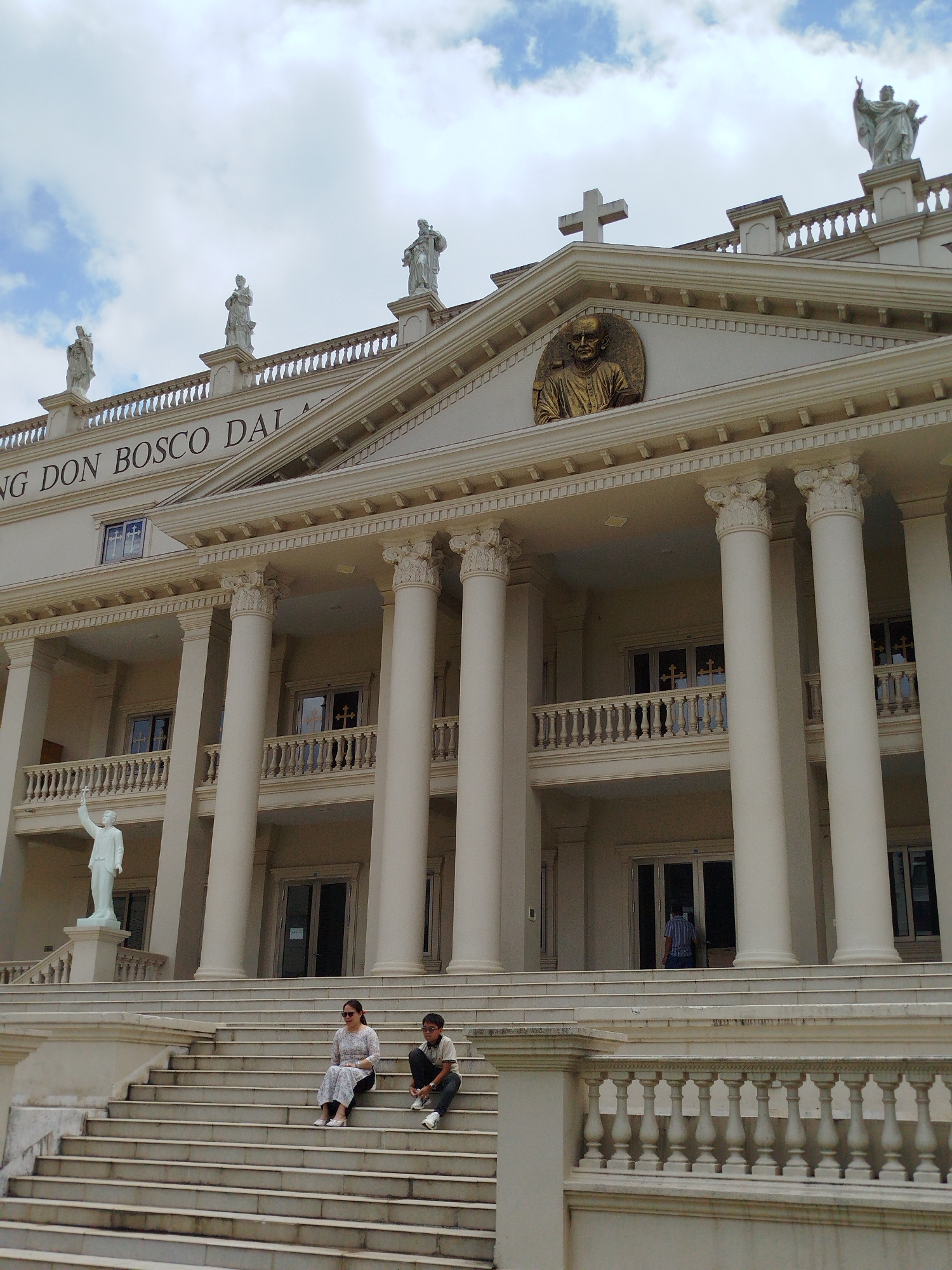 The image depicts the grand facade of Don Bosco Da Lat, a Catholic church located in Da Lat, Vietnam. The building is a stately structure with classical architectural elements, including tall white columns, a pediment, and statues. The name "NG DON BOSCO DALAT" is inscribed above the columns. Statues of religious figures are positioned along the roofline and on the grounds. A large cross adorns the peak of the pediment. A golden relief of Don Bosco is visible just below the cross.
Two individuals, an adult woman and a teenage boy, are seated on the steps leading up to the building. A third person is partially visible entering the building's doorway. The weather appears cloudy. The scene is captured from a low angle, emphasizing the building's scale.