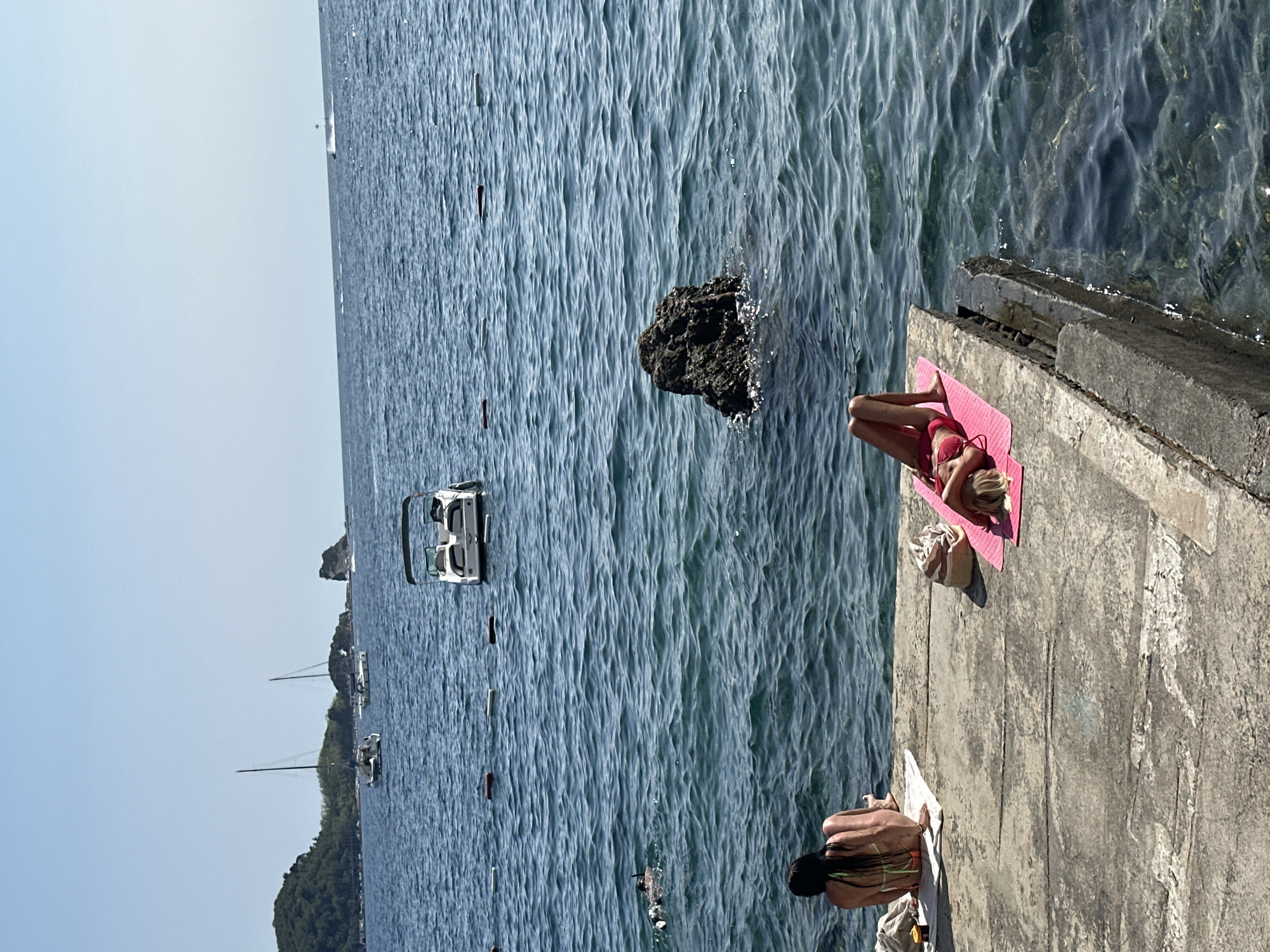 The image depicts a sunny day at the coast of Budva, Montenegro, likely near a beach or marina. In the foreground, two women are sunbathing on a concrete platform by the sea. One woman, with long dark hair, is seated with her back to the camera, wearing a bikini. The other woman, with blonde hair, is lying on her back on a pink mat, also in a bikini. Several bags and towels are scattered around them.
The sea is a deep blue, with gentle waves. In the middle ground, a white motorboat is anchored, and several buoys mark a swimming area. Further out, two sailboats are visible, and a person is swimming. In the background, a tree-covered island or peninsula rises from the sea, and a distant rocky outcrop is also visible. The sky is clear and blue. The scene conveys a relaxed atmosphere of a summer day at the seaside.