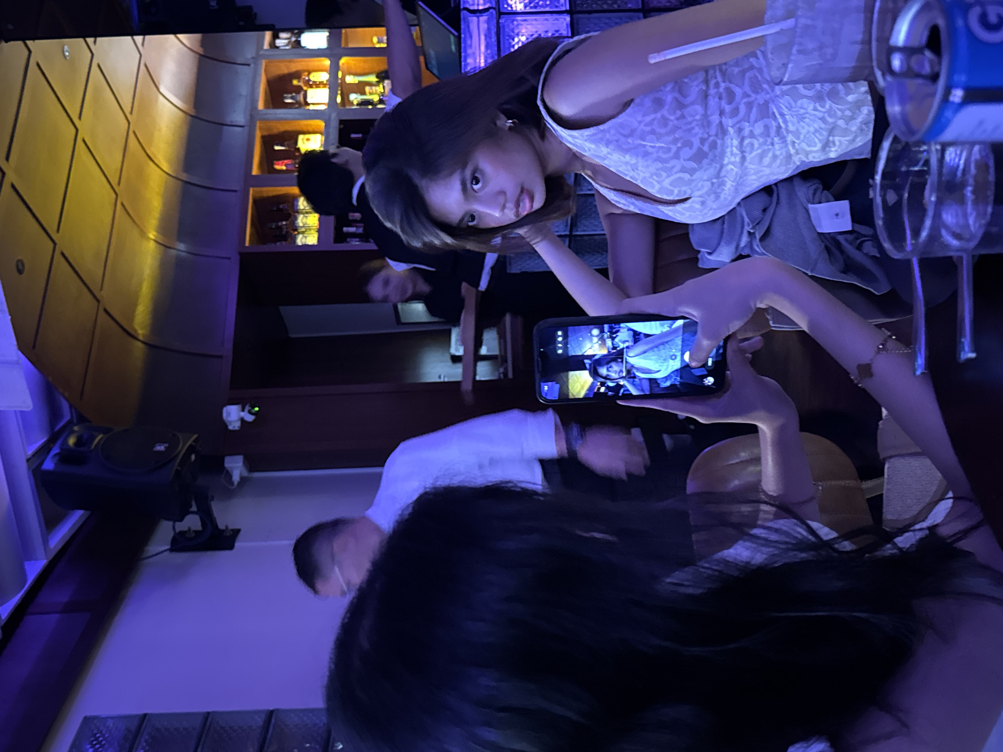 In a dimly lit establishment in Davao City, Philippines, a woman with dark hair, wearing a white lace top, poses for a photo on a smartphone. She is seated, with her right arm resting on a brown cushioned surface. Her gaze is directed towards the camera of the smartphone being held by another person, whose hands and arms are visible, framing the phone. The smartphone screen displays a mirrored image of the woman. The background reveals a bar area with shelves stocked with bottles, illuminated by warm lighting. Other individuals are discernible in the background, including a man in a white collared shirt and glasses, and a woman in a black uniform. The overall ambiance suggests a social gathering or a night out at a bar or restaurant.