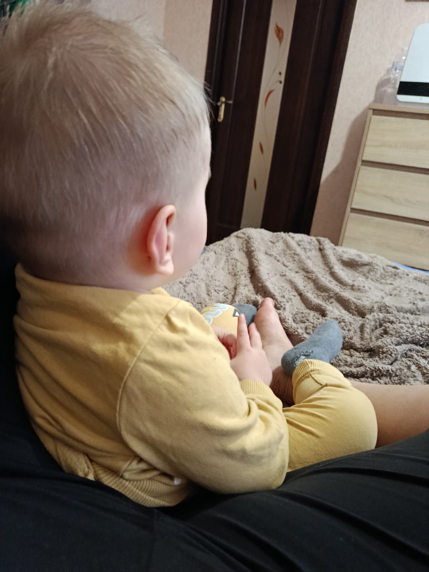 The image captures a close-up, high-angle shot from behind a baby, seemingly sitting on someone's lap, in what appears to be an indoor setting. The baby, with light blonde hair, is wearing a yellow long-sleeved top and matching yellow pants. Their head is turned slightly to the right, revealing their ear. One foot is bare, and the other is wearing a grey sock. The baby's hands are interacting with their bare foot, possibly playing or exploring.
In the background, a crumpled brown textured blanket or throw is visible, suggesting a bed or couch. To the right, part of a light-colored wooden dresser with horizontal drawers can be seen, and on top of it, a white electronic device and a clear glass object. Further to the left, a dark wooden door with decorative lighter-colored panels and a handle is visible. The walls are a light, possibly peach or beige, color. The lighting suggests an indoor setting, likely during the day or with artificial light. There is no visible text in the image. The scene conveys a sense of quiet observation or play.