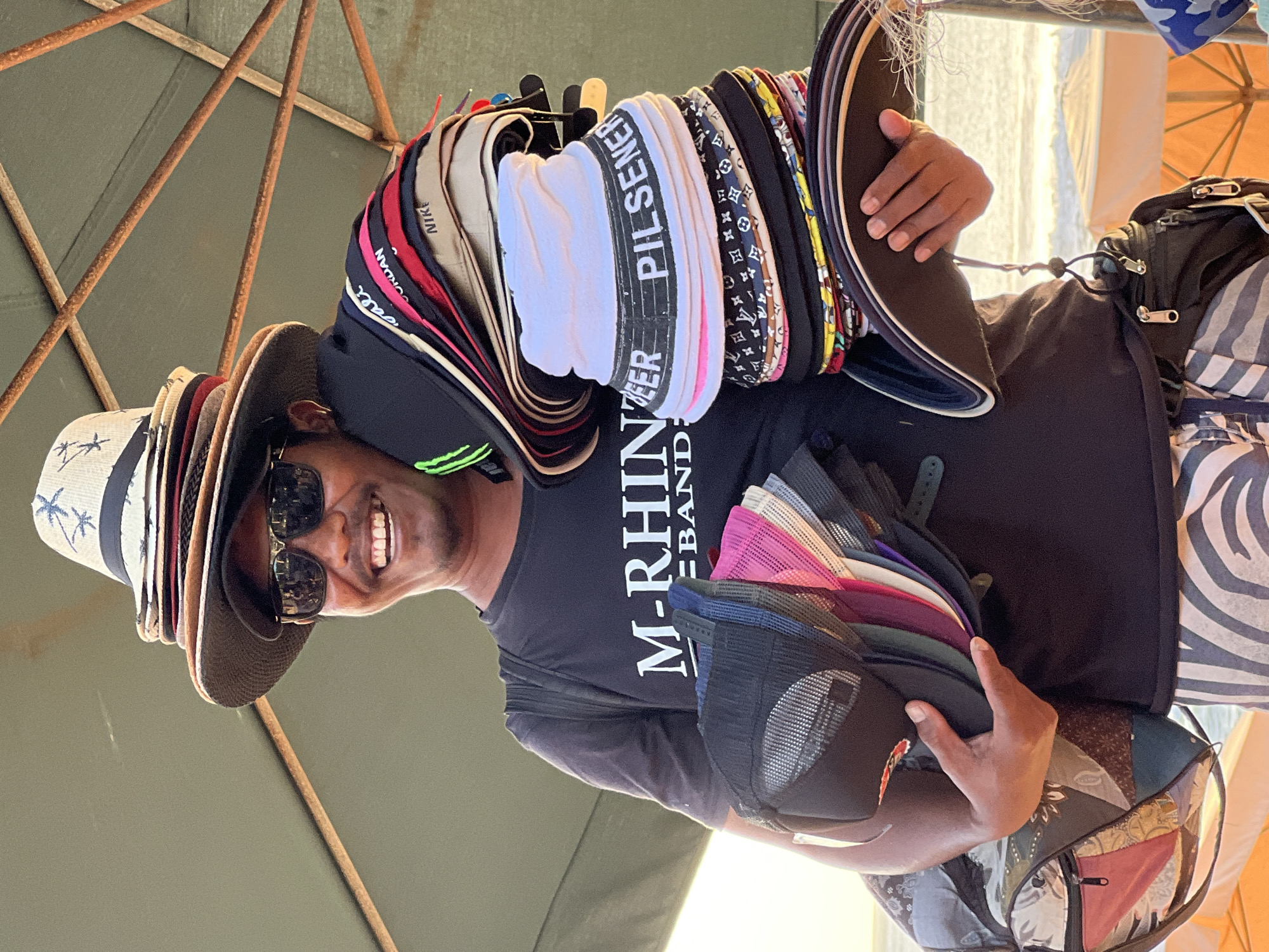 In Kuta, Indonesia, a man stands under a green umbrella, offering hats for sale. He wears a black t-shirt with the words "M-RHINO BAND" in white and dark sunglasses. A stack of fedora hats is balanced on his head. In his arms, he holds a collection of baseball caps and bucket hats. Some of the caps are colorful, with brands like "NIKE" and "JORDAN" visible. A duffel bag with a patchwork design rests on his left side. The background shows a beach with gentle waves and sun umbrellas. The man is smiling, presenting his merchandise to potential customers.