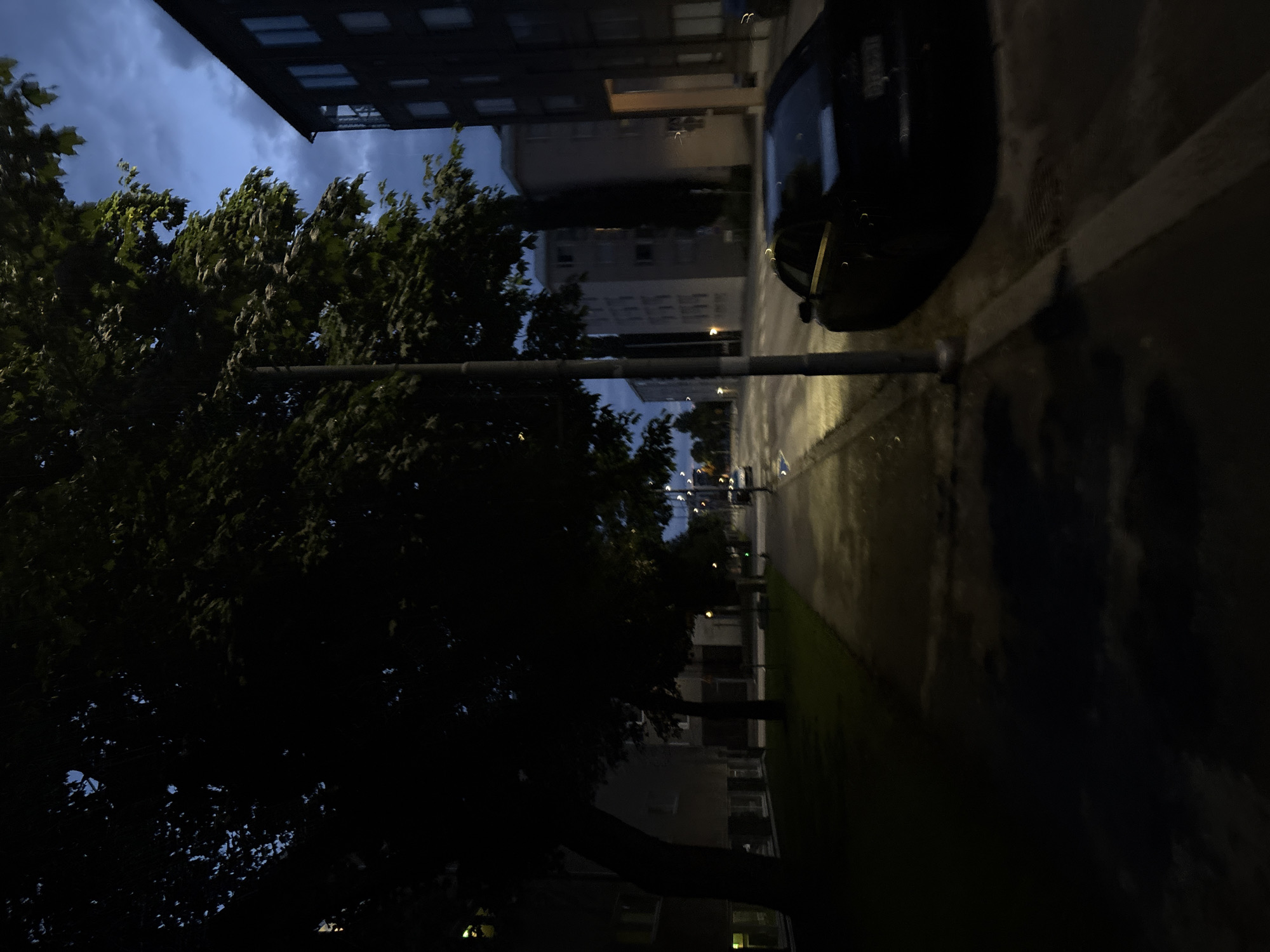 The image depicts an evening scene on a street in Pori, Finland. A dark-colored sedan is parked on the right side of the street, facing away from the camera. The street itself is paved, with a sidewalk to the left, bordered by a grassy area. Trees with dense foliage line the sidewalk, casting long shadows. Several apartment buildings are visible in the background, some appearing darker and more modern than others. The sky is a deep blue, with some clouds, indicating twilight or early night. A lamppost stands near the parked car, its light casting a glow on the pavement. The overall impression is one of a quiet urban residential area during the evening.