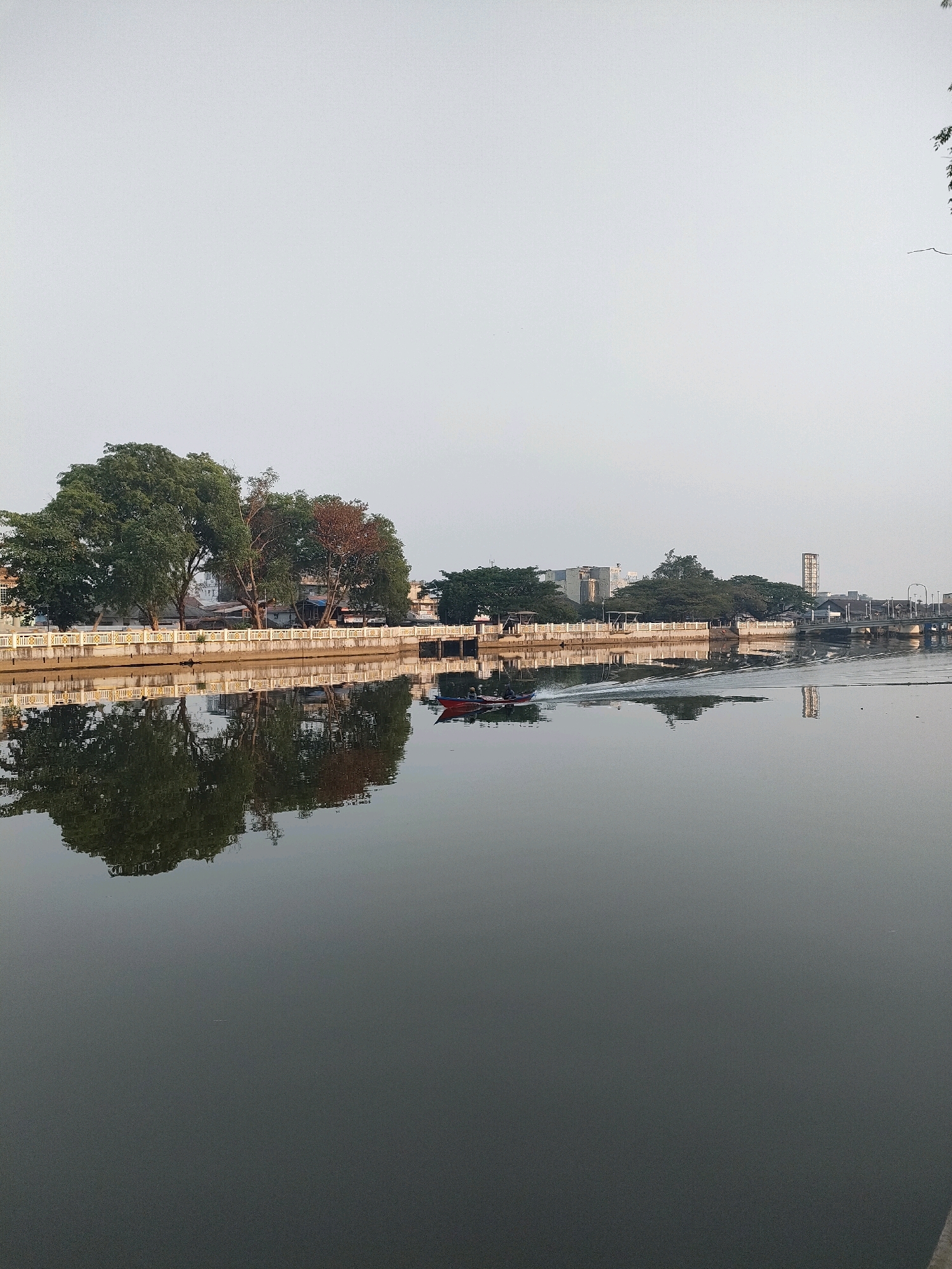 An exterior, vertical, eye-level shot captures a serene canal in Banjarmasin, Indonesia, under a pale, overcast sky. The canal's still, dark water reflects the opposite bank, where a white, ornate railing lines the promenade. Behind the railing, lush green trees, their foliage dense, stand tall. Several buildings, some appearing residential and others commercial, are visible beyond the trees, with a prominent clock tower or water tower standing out in the distance. A narrow, red boat with two occupants glides across the canal, leaving a white wake that disrupts the perfect reflection. The scene evokes a sense of calm and everyday life in the city.