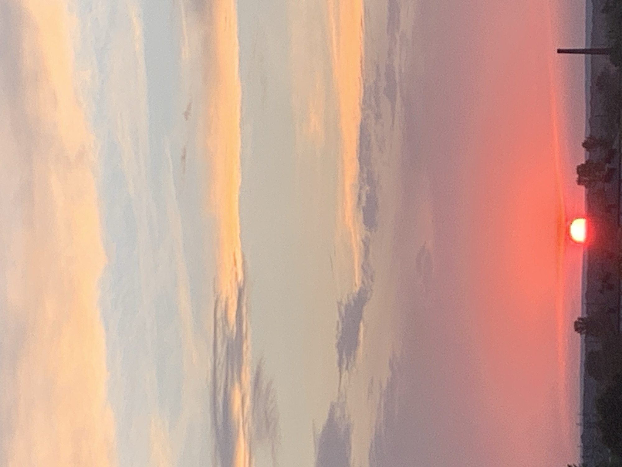 The image shows a sunset over a city. The sky is filled with clouds in shades of pink, orange, and blue. The sun is setting on the horizon, casting a warm glow over the scene. In the foreground, there is a silhouette of trees and buildings. The image appears to have been taken in Kolomyia, Ukraine, based on the context.