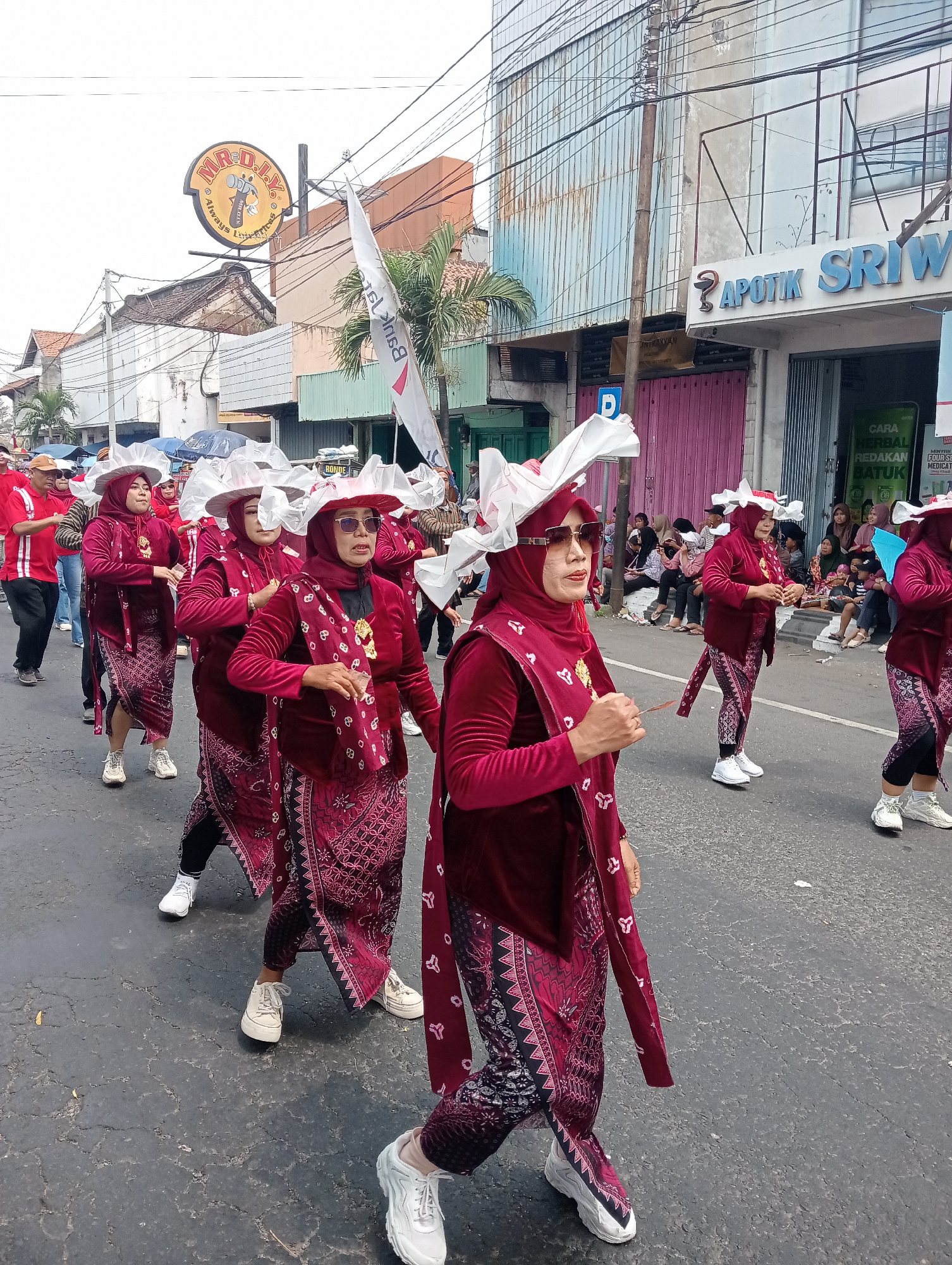The image shows a parade in the city of Demak, Indonesia, likely on Jalan Kyai Singkil, based on the presence of a MR. DIY store sign which is common in Indonesian cities and the architectural style of the buildings. Several women are walking in a procession, dressed in matching maroon outfits with intricate patterned skirts and headscarves. They are also wearing large, white, ruffled hats that create a dramatic visual effect. Some of the women are wearing sunglasses. The women are marching on a paved road, and there are spectators sitting on the side of the road watching the parade. In the background, there are buildings, including a store labeled "APOTIK SRIW" (likely Apotek Sriwijaya), a common pharmacy chain in Indonesia. A white flag with writing is also visible. The overall scene suggests a community event or cultural celebration.