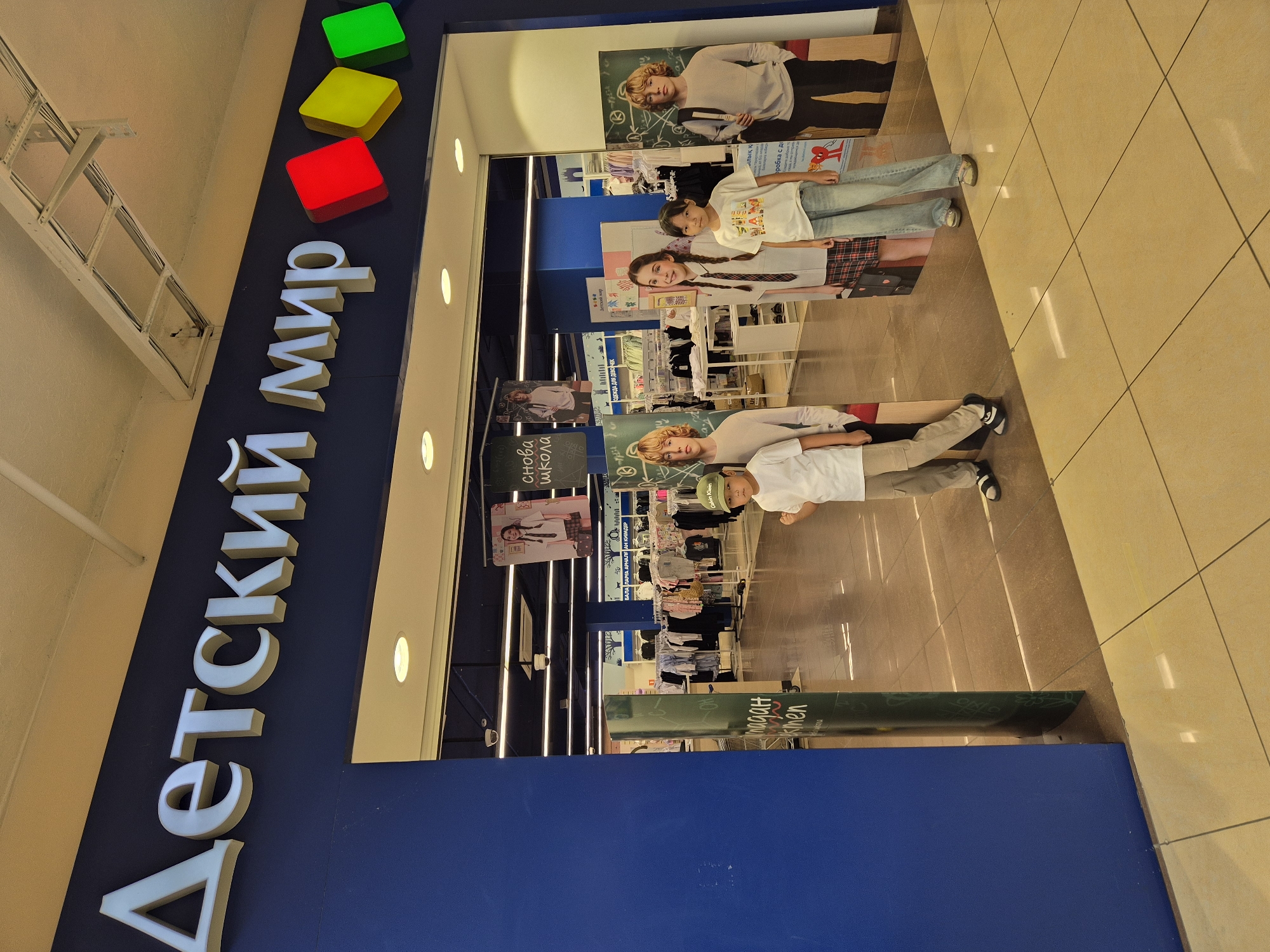 The image shows the entrance to a "Detsky Mir" (Children's World) store. The store's name is displayed in large, white, three-dimensional letters on a dark blue facade. To the right of the name are three colored squares: red, yellow, and green, which appear to be part of the store's branding.
In the foreground, standing in the entrance, are three children. The child on the left is a boy wearing a white t-shirt and khaki pants, with a green baseball cap. He is facing forward with his right fist raised. Next to him is a younger child, a girl, wearing a white t-shirt with graphics and blue jeans. She is looking towards the camera. To the right of her is a boy, also wearing a light-colored t-shirt and dark pants, with a backpack slung over his shoulder. He is looking to his right.
Behind the children, inside the store, racks of children's clothing are visible. There are also large cardboard cutouts of children in school uniforms, advertising back-to-school items. Posters with images of children and text are hanging from the ceiling. The floor inside the store appears to be tiled with light-colored square tiles.
The overall environment suggests a retail store specializing in children's products, likely a department store. The presence of school-themed advertising indicates it might be the back-to-school shopping season. The image does not provide any specific geographical markers that would definitively place it in Kokshetau, Kazakhstan, beyond the assumption based on the language on the signage which is Russian.