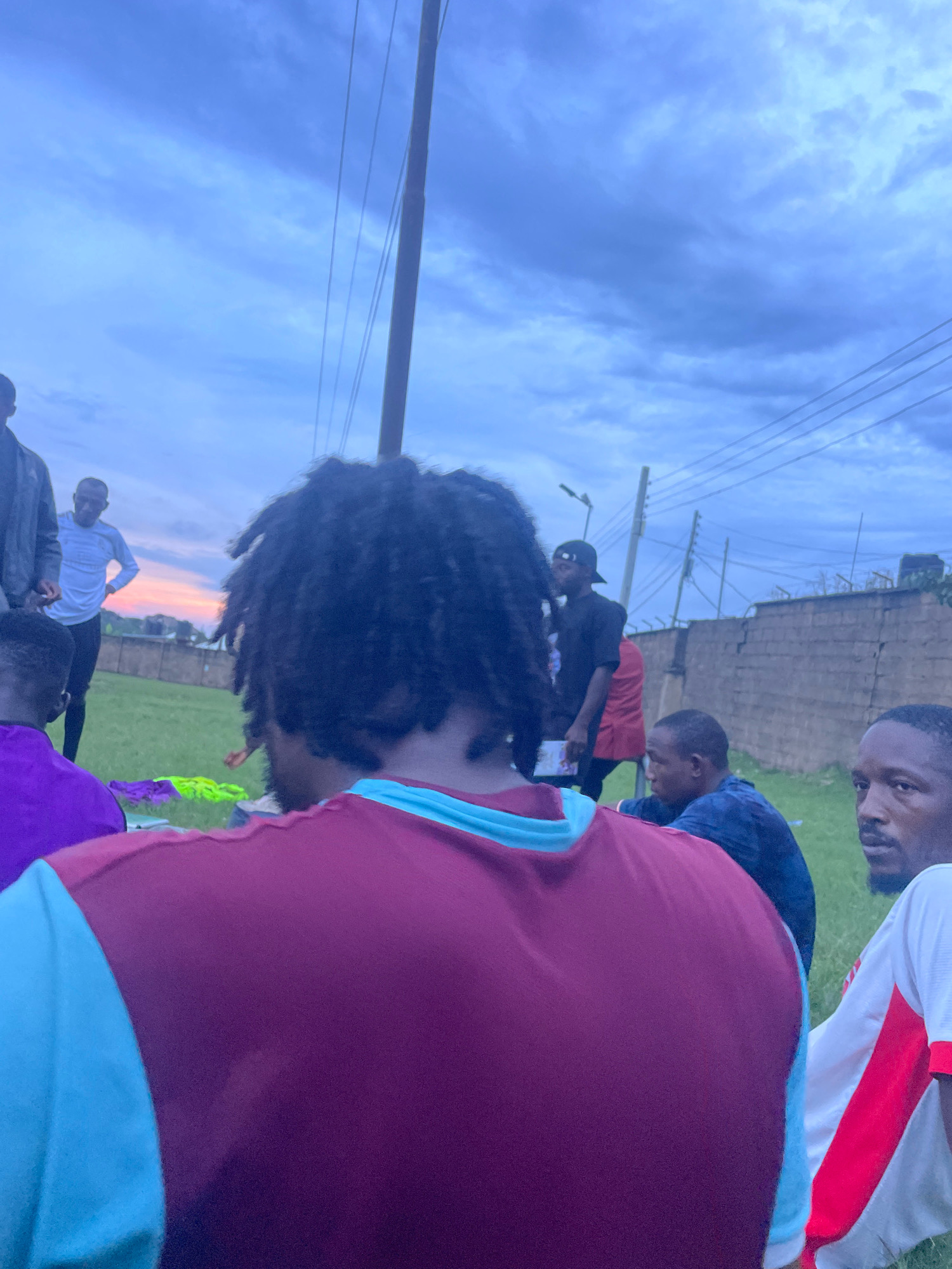 In the city of Jos, Nigeria, at dusk, a group of individuals are gathered outdoors in a grassy area. The sky is overcast with dramatic clouds, transitioning into hues of orange and pink near the horizon. Power lines are visible crisscrossing the sky, supported by a tall pole and other utility structures. In the foreground, the back of a person's head with dreadlocks is prominent, wearing a maroon and teal shirt. To their left, another person in a purple shirt is partially visible. Further back, a group of men are conversing and observing. One man in a white long-sleeved shirt stands with his hands on his hips, facing the sunset. Another man in a black t-shirt and baseball cap looks towards the left. A brick wall with a metal fence runs along the background, with some buildings and trees beyond. The overall impression is a casual outdoor gathering during twilight.