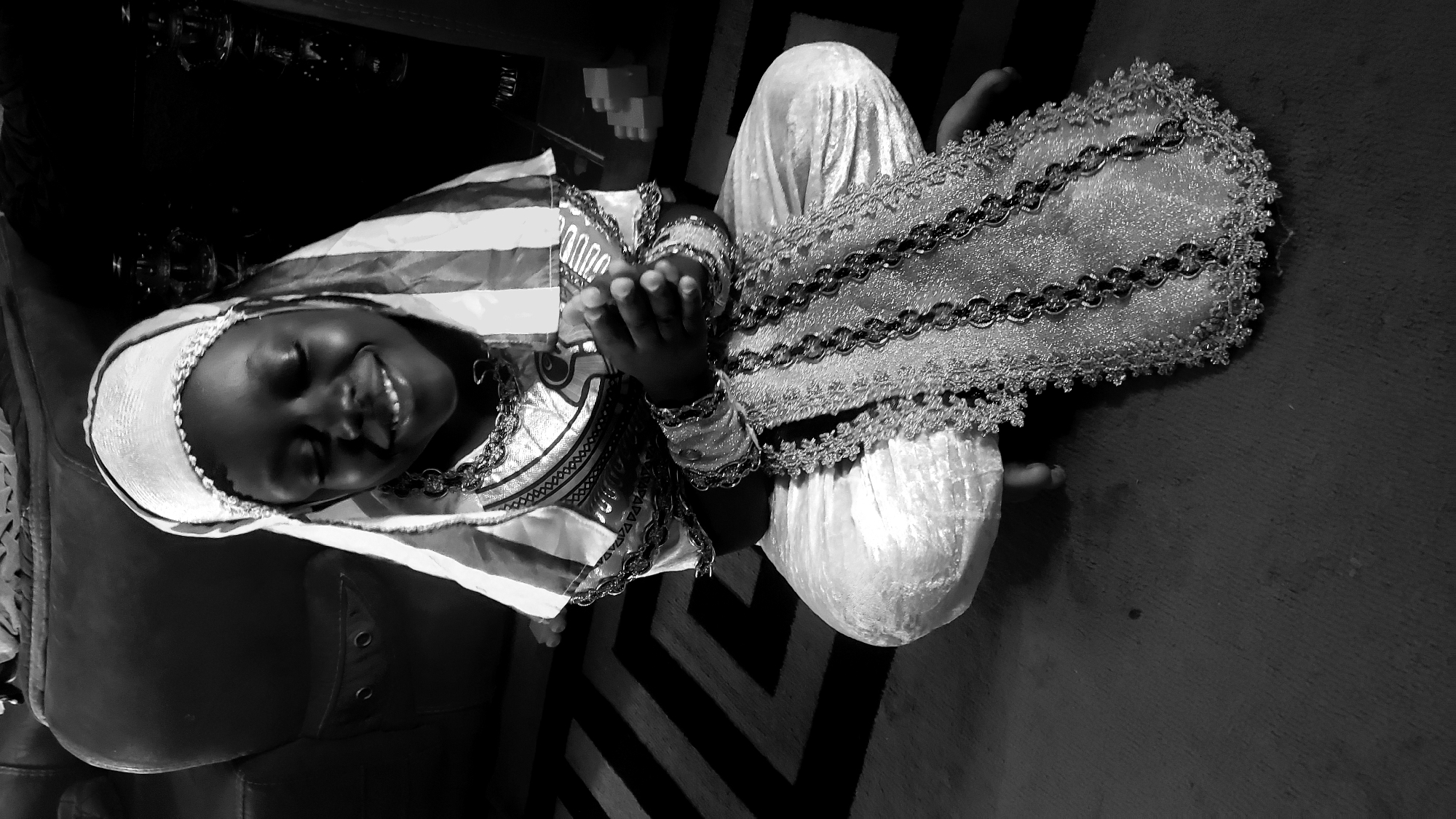 A black and white, top-down, high-angle shot shows a young Black person dressed in traditional Nigerian attire, sitting cross-legged on a carpeted floor. The person is smiling with their eyes closed, appearing content. They are wearing a white headscarf with a beaded trim, a white and striped garment with a decorative chain necklace, and what appear to be bangles on their wrists. The outfit is elaborate and appears to be made of shiny, textured fabric. The person is holding a decorative, rectangular piece of fabric adorned with intricate silver and black embroidery and beading. The setting is indoors, with a dark, possibly leather couch visible on the left and a patterned carpet in the background. The overall mood is joyous and celebratory, possibly indicating a special occasion. The lighting suggests it is daytime, with no indication of specific weather.