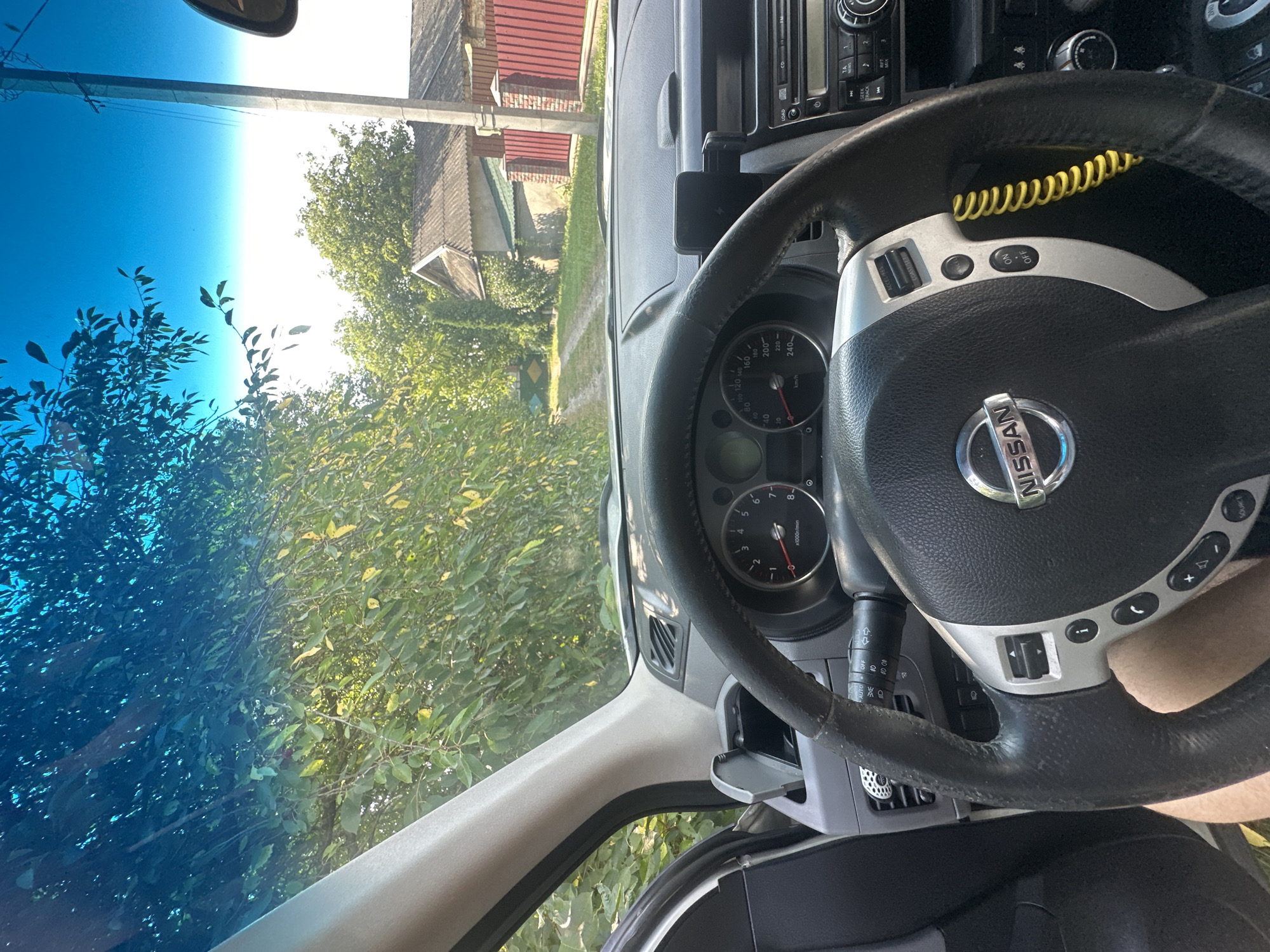 The image is taken from the interior of a car, looking out through the windshield. The dashboard and steering wheel of a Nissan vehicle are visible in the foreground. The steering wheel has various controls on it, including buttons for cruise control and audio. A phone mount is attached to the dashboard. Outside the car, a dirt road leads into a rural landscape. Trees and houses with tiled roofs are visible. The sky is bright blue, with some leaves from trees casting shadows. The scene appears to be a sunny day in a rural area, possibly a village or countryside location. Given the context of Shatava, Ukraine, the image likely depicts a road within or on the outskirts of this Ukrainian town.
