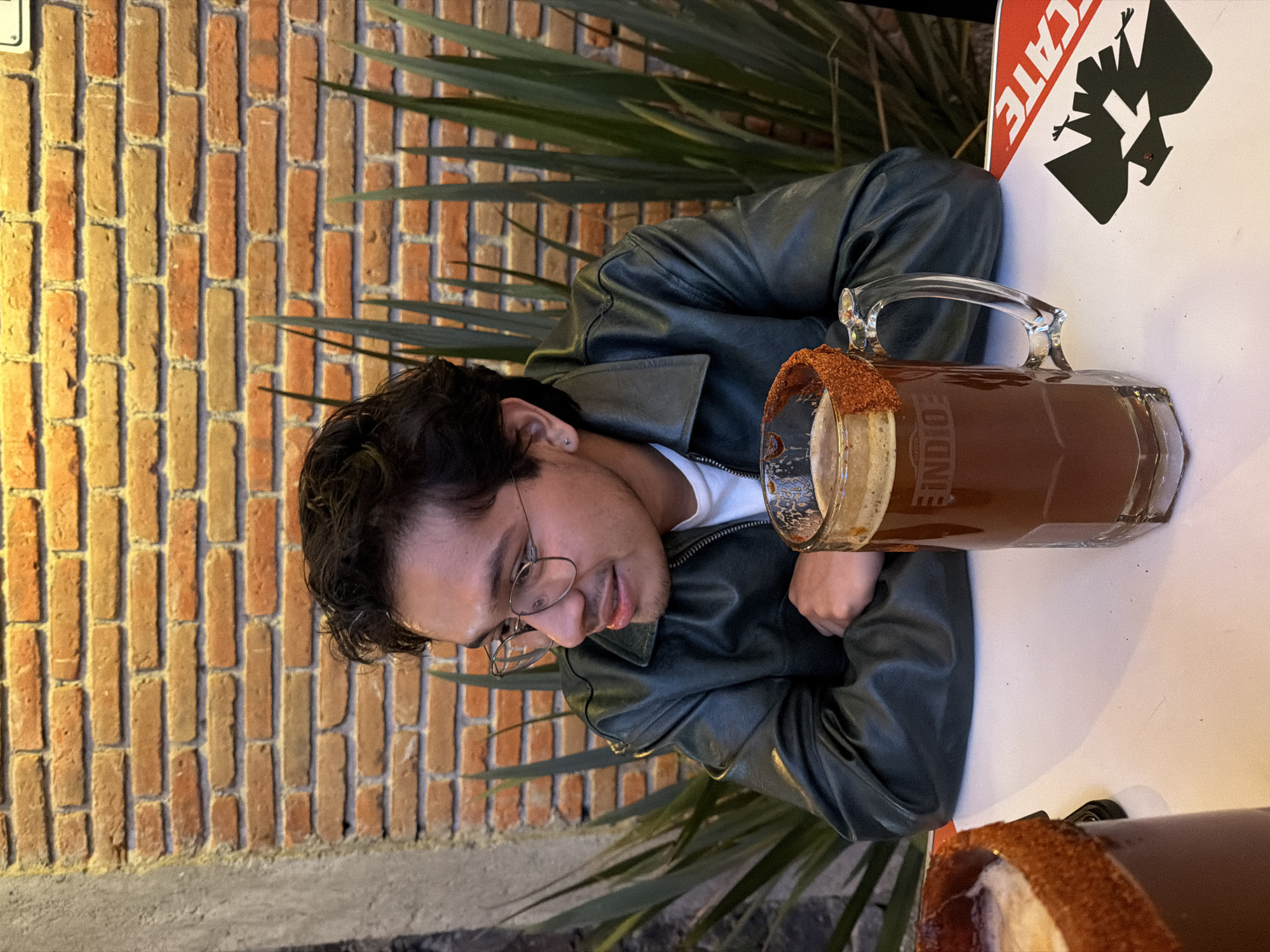 A young man in glasses and a leather jacket sits at a white table, upon which sits a large beer mug, rimmed with a reddish-brown powder, and filled with a dark liquid. The man is looking down at the mug. Behind him is a brick wall, with tall, spiky green plants in front of it. A sign, partially visible, reads "CATE." The beer mug is branded with the words "INDIO CERVEZA." The location is San Andrés Cholula, Mexico.