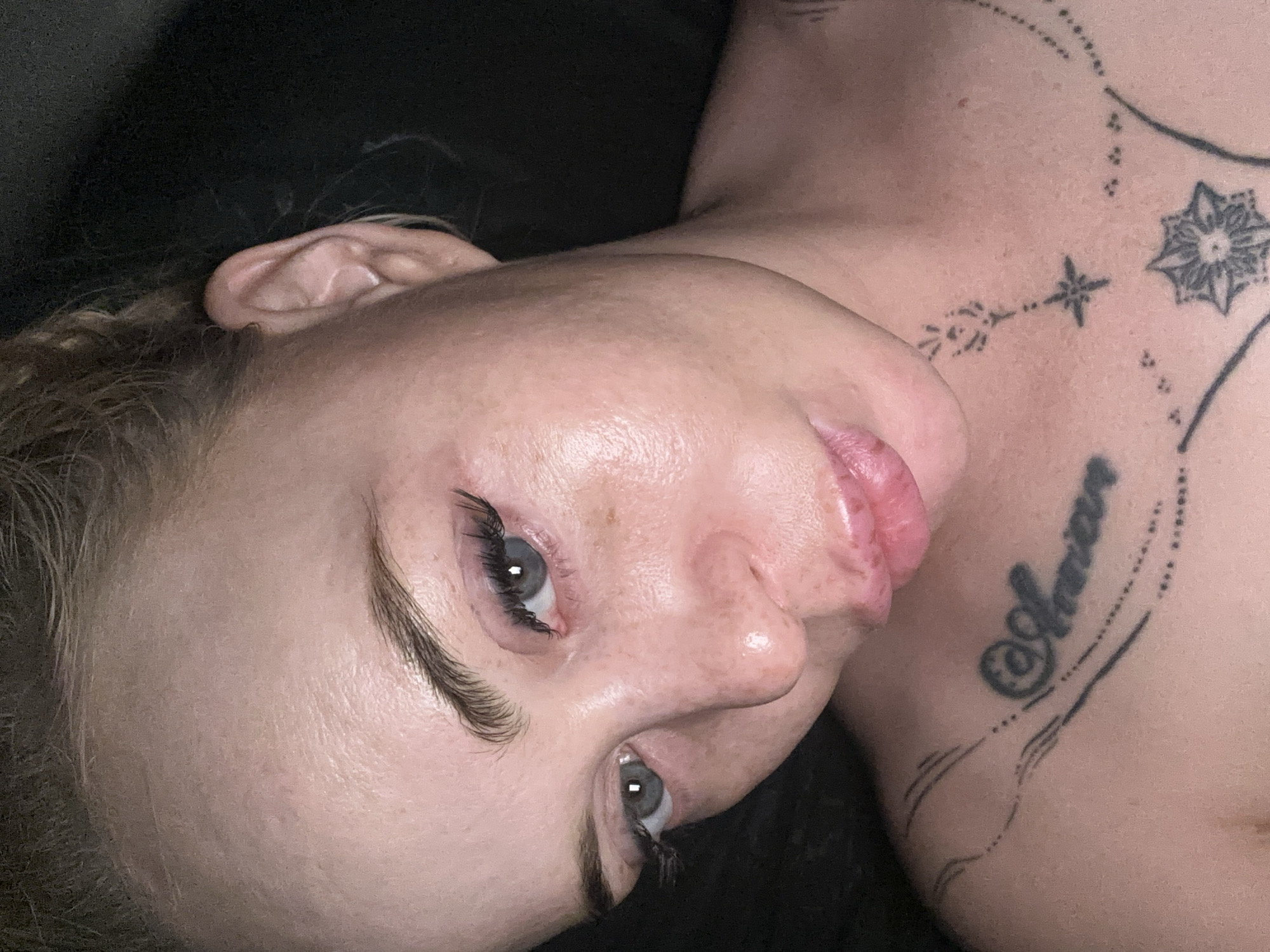A close-up, head and shoulders shot shows a light-skinned person with blonde hair pulled back. They are looking down and to their left. The person is wearing makeup, including dark eyeliner and mascara. The person has tattoos on their chest and neck. The background is dark and out of focus. The image was taken in an unknown location.