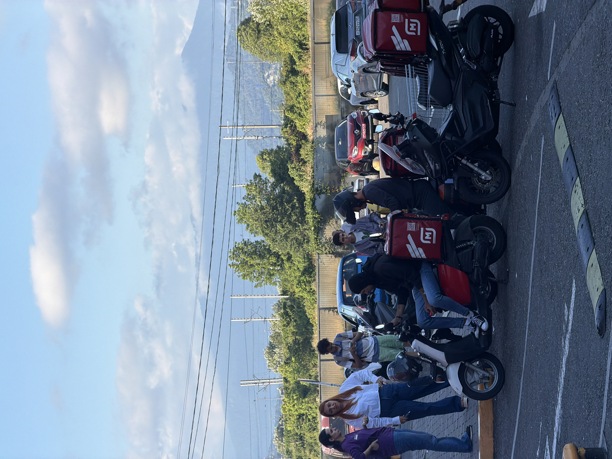 The image depicts a group of people gathered around several motorcycles and cars parked on a paved surface. The scene appears to be outdoors, with a backdrop of lush green trees and rolling hills or mountains under a bright, partially cloudy sky. The lighting suggests it is daytime, likely late afternoon or early evening given the shadows.
The subjects include several individuals, some of whom are wearing casual clothing and others appear to be wearing delivery uniforms with red insulated bags on the motorcycles. The motorcycles are equipped with red delivery boxes on the back, featuring a white logo that resembles the letter 'M' with a wing-like shape. One car, a red Renault, has a license plate that reads "PRODAETSIA," which is Russian for "FOR SALE." Another car, a silver SUV, has a license plate with Cyrillic characters.
The people are interacting with each other and the vehicles, with some standing and others seated on the motorcycles. The overall atmosphere seems relaxed, with no overt signs of distress or urgency. The presence of multiple delivery vehicles and the gathering of people might indicate a meeting point for delivery drivers, a break in their workday, or perhaps a promotional event. The setting includes power lines and poles, suggesting an urban or suburban environment, consistent with the provided location context of Sirius, Russia.