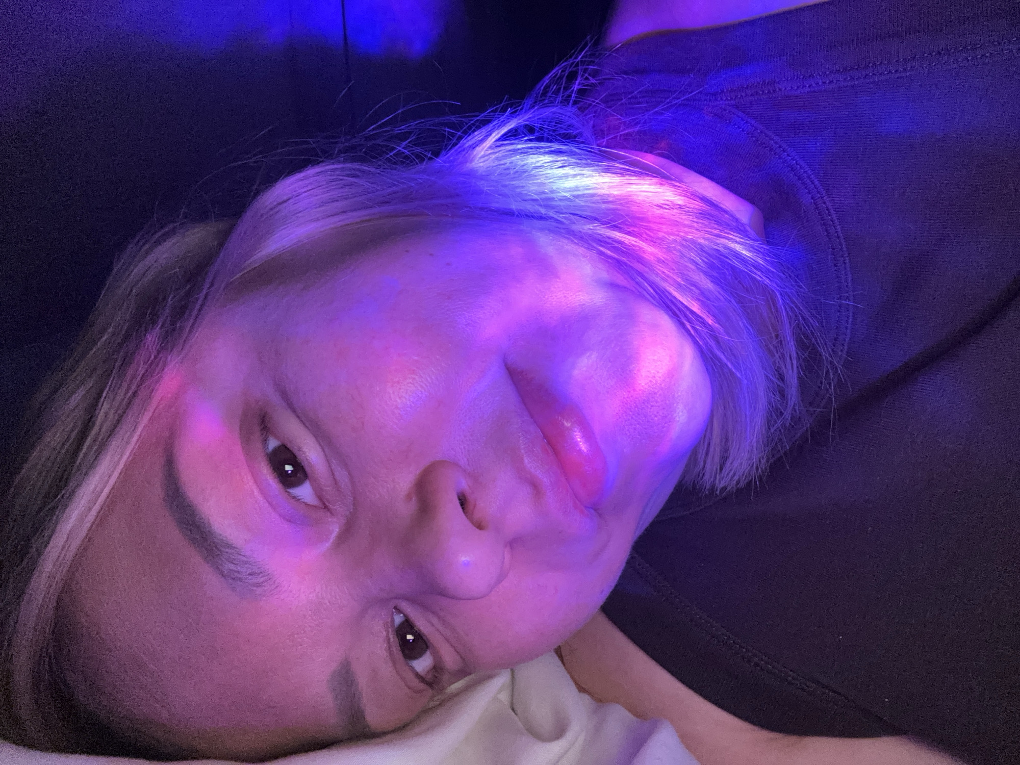 A close-up, angled portrait shows a person lying on a white surface, likely a pillow or bedding. The person has fair skin, brown eyes, and light blonde hair with darker roots. Their face is illuminated by a combination of pink and blue light, with the blue light predominating in the upper portion of the head and hair. The person's facial expression suggests a playful or slightly seductive mood. They are wearing a dark-colored top, visible in the upper right corner of the frame. The background is dark and indistinct. There is no information in the image to suggest a location in Izhevsk, Russia.