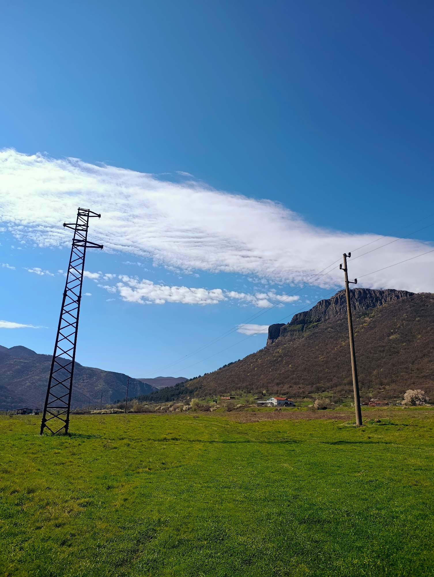 The image depicts a rural landscape in Shiroko pole, Bulgaria, under a bright blue sky with scattered white clouds. In the foreground, there's a green field with two types of electric power transmission towers: a tall, steel lattice tower to the left and a shorter, wooden pole structure to the right. Wires connecting the poles are visible. In the background, there is a mountain range with sparse vegetation, and a few buildings are nestled at its base. The light suggests a sunny day with clear visibility.