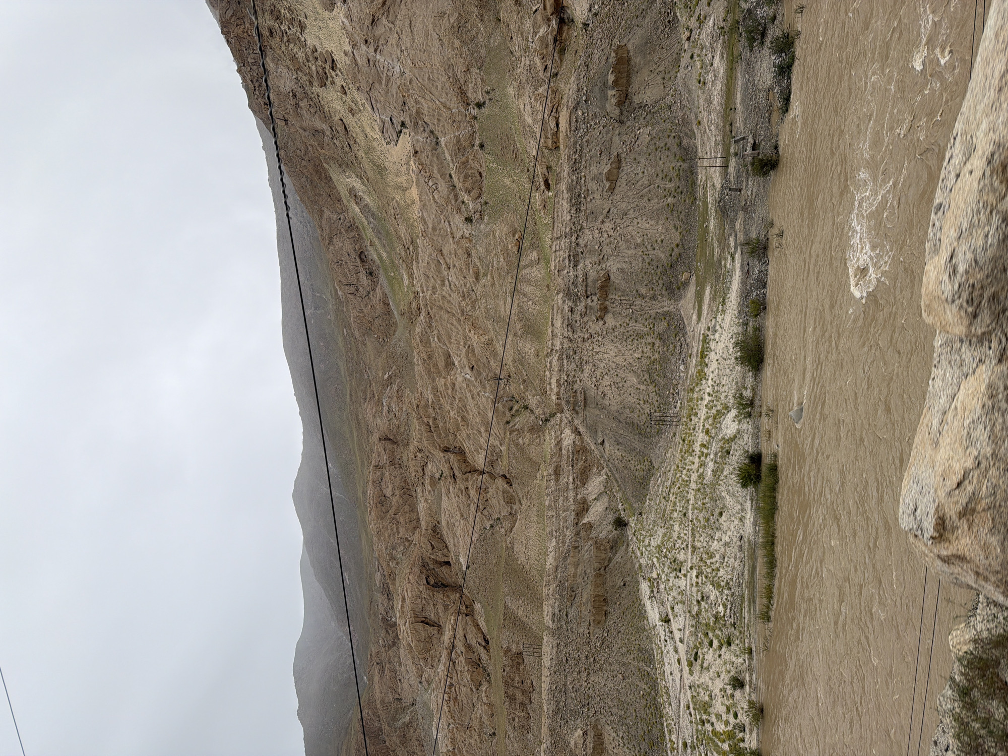 The image depicts a wide, muddy river flowing through a valley with steep, barren, rocky mountains on either side. The sky is overcast and gray. Power lines are visible stretched across the frame, running horizontally in the upper part of the image and diagonally in the lower part. The river appears to be at a high level, with turbulent water and white foam. Sparse green vegetation grows along the riverbanks and on the lower slopes of the mountains. The scene is desolate and rugged, indicative of a mountainous, possibly arid region. There are no people or animals visible in the image. The location, as stated, is Kungyamdo, India, which is a village in the Zanskar region of Ladakh. This region is known for its dramatic mountainous terrain and rivers, often influenced by monsoon rains or snowmelt, which can cause rivers to swell and become muddy.