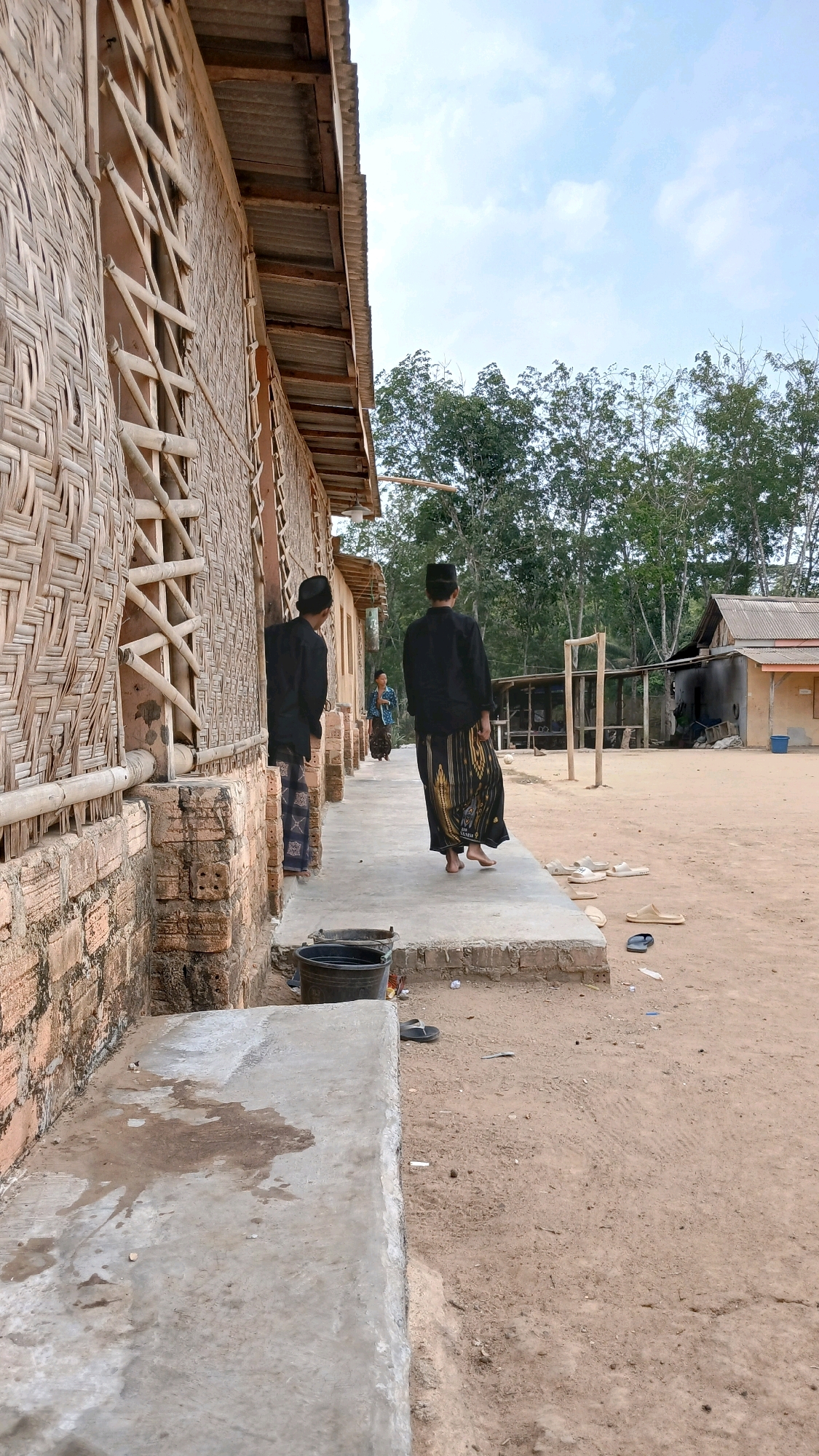 The image depicts an outdoor scene in Adi Luhur, Indonesia. In the foreground, a dirt path runs alongside a building with walls constructed from woven bamboo and brick. Two individuals, dressed in black tops and sarongs, are walking away from the camera on a concrete walkway that borders the building. One of them is barefoot. Further down the path, a third person, a woman in a blue and patterned outfit and sarong, is also walking away from the camera. In the background, several other simple structures, including one with a corrugated metal roof, are visible. Trees with dense foliage form a backdrop against a light blue sky with scattered clouds. Scattered on the ground are several pairs of sandals, some buckets, and debris. The overall impression is of a rural or semi-rural residential area.