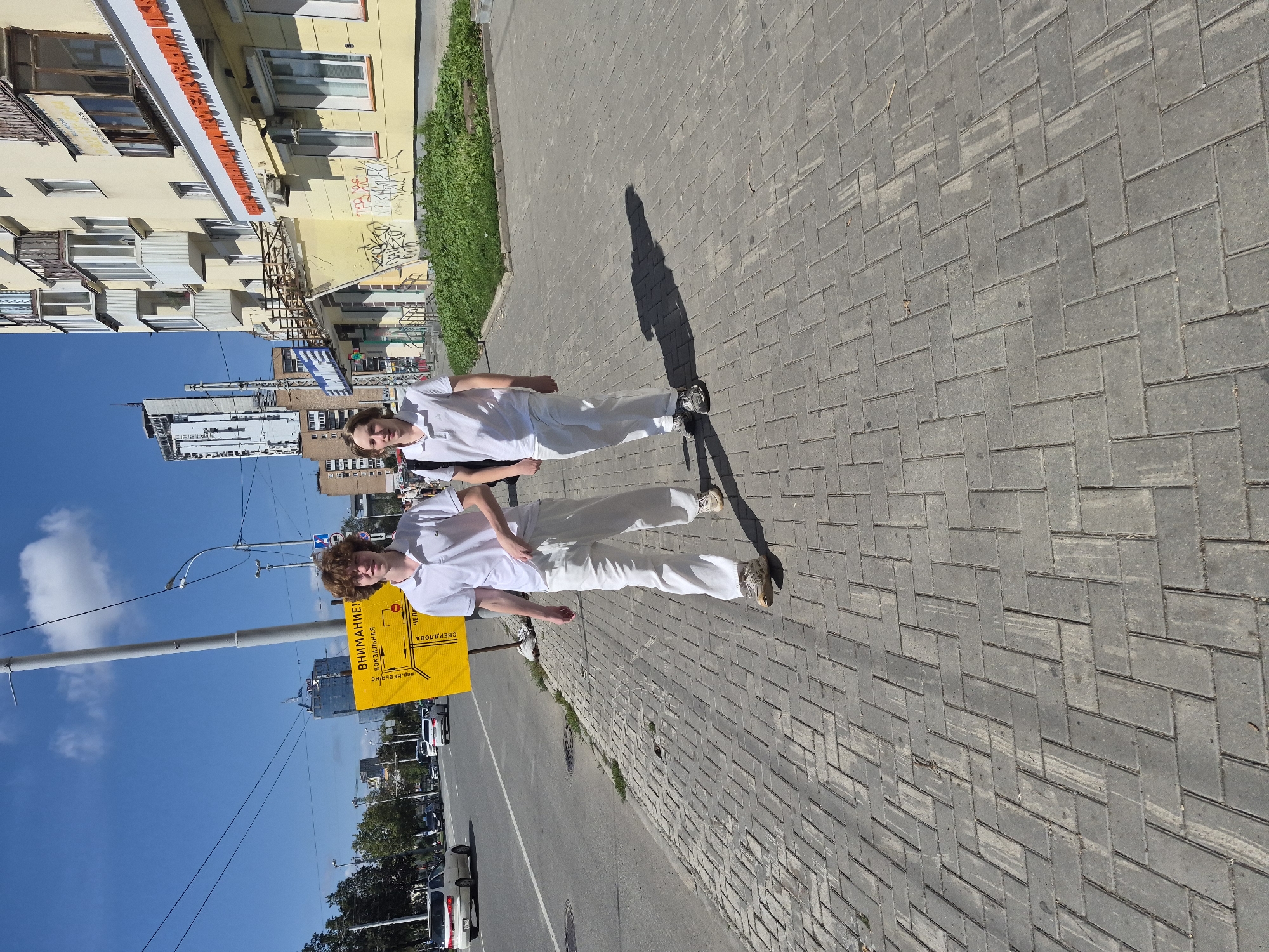 Two young men are walking on a city sidewalk in Yekaterinburg, Russia, on a sunny day. The man on the left has curly brown hair and is wearing a white polo shirt and white pants. The man on the right has lighter brown hair and is wearing a white polo shirt and white pants. They are both wearing sneakers. The sidewalk is paved with gray interlocking bricks. To the left of the sidewalk is a road with cars driving on it. To the right of the sidewalk is a yellow sign with Russian text and a map, and behind that, a modern building with balconies and windows. Further in the background, there are other city buildings and trees. The sky is blue with some clouds.