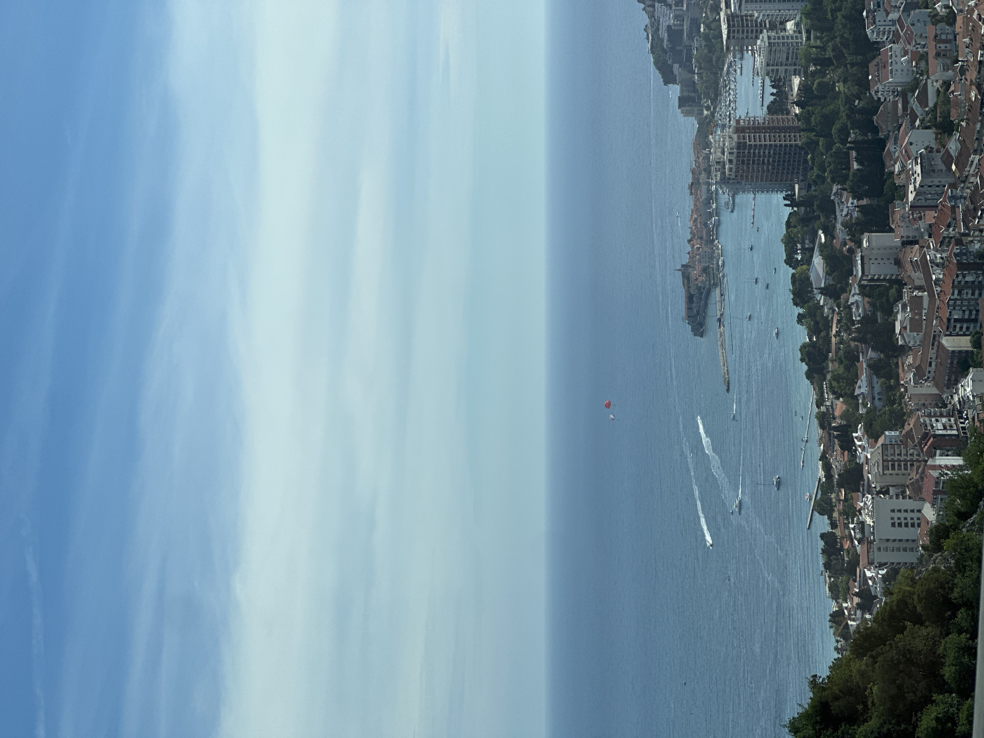This is a high-angle shot of a coastal city in Vrela, Montenegro. The sea occupies the top half of the image, with a clear horizon line. In the water, two boats are visible with white wakes trailing behind them, and one sailboat is anchored. Further out, a red parachute is visible in the sky, attached to a person or object that is not clearly discernible. The city spans the lower half of the image, characterized by buildings with red-tiled roofs and some taller, modern structures. Green trees are interspersed throughout the urban landscape. The sky is a pale blue with thin, wispy clouds. A portion of a forested, hilly area is visible in the lower left corner of the frame.