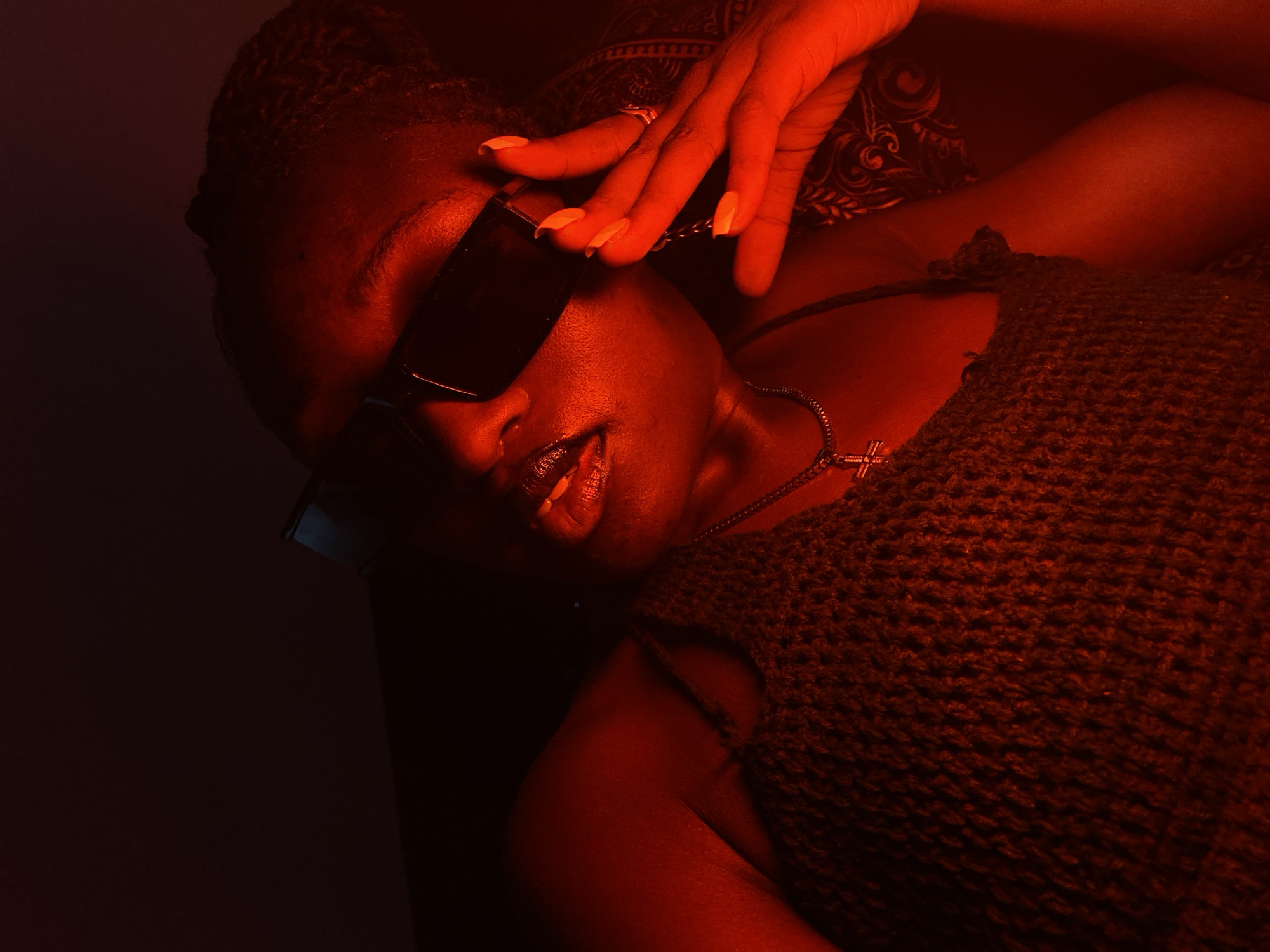 In a dimly lit environment with strong red lighting, a person with dark skin, wearing sunglasses, a knitted top, and a cross necklace, poses for a portrait. The person's right hand is raised, touching their face. The background is dark and indistinct, with some patterned fabric visible on the right. The scene does not contain any elements that would allow for the determination of its location within Bar Jirgi Summa, Nigeria.