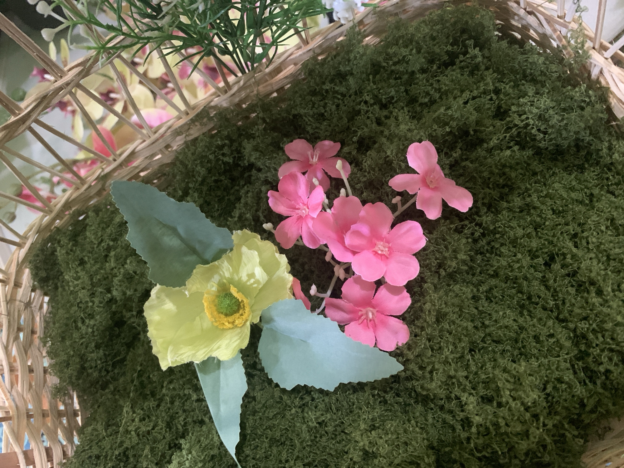 A close-up, high-angle shot shows a decorative arrangement inside a woven basket. The basket is filled with a dense, green, moss-like material. Scattered on top of the moss are several artificial flowers: one pale yellow poppy with a green stamen and two light blue leaves, and a cluster of five small pink flowers. The basket is positioned within a room, suggested by the blurred background which includes other decorative elements and what appear to be other artificial floral arrangements. There are no people or animals present in the image. The setting is likely an indoor decorative display, possibly for a home or event, and there is no discernible connection to Kuala Pembuang, Indonesia.
