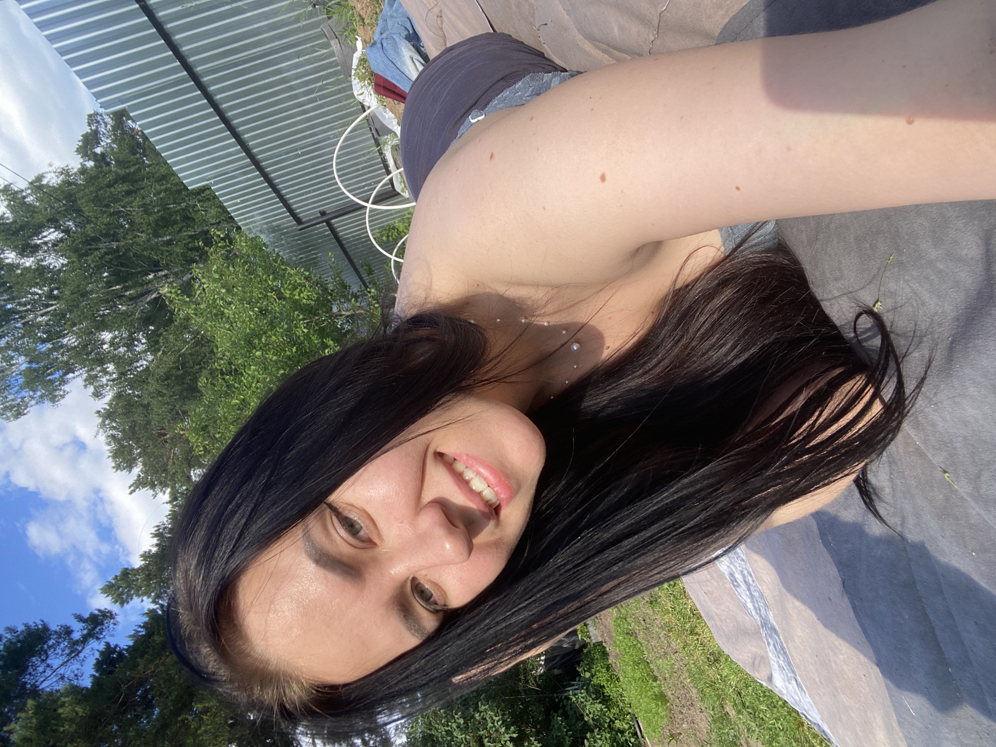 A young adult female with long, straight, dark hair is depicted lying prone on a light-colored blanket or mat in an outdoor setting, likely a backyard or garden area, in the city of Chaikovsky, Russia. She is positioned with her head and upper torso angled towards the camera, smiling broadly and revealing white teeth. Her eyes are greenish-brown with black winged eyeliner. She is wearing a gray sleeveless top and a delicate necklace featuring multiple small, pearl-like drops. Partial denim shorts are visible on her lower body. The scene is brightly lit by direct sunlight, casting distinct shadows on her arm and the ground cover. In the background, green grass and various trees (including both coniferous and deciduous types) are visible. A tall, corrugated silver metal fence with black horizontal supports runs across the mid-ground. Near the fence, white arched structures (possibly garden hoops) and crumpled blue fabric are present on the ground.