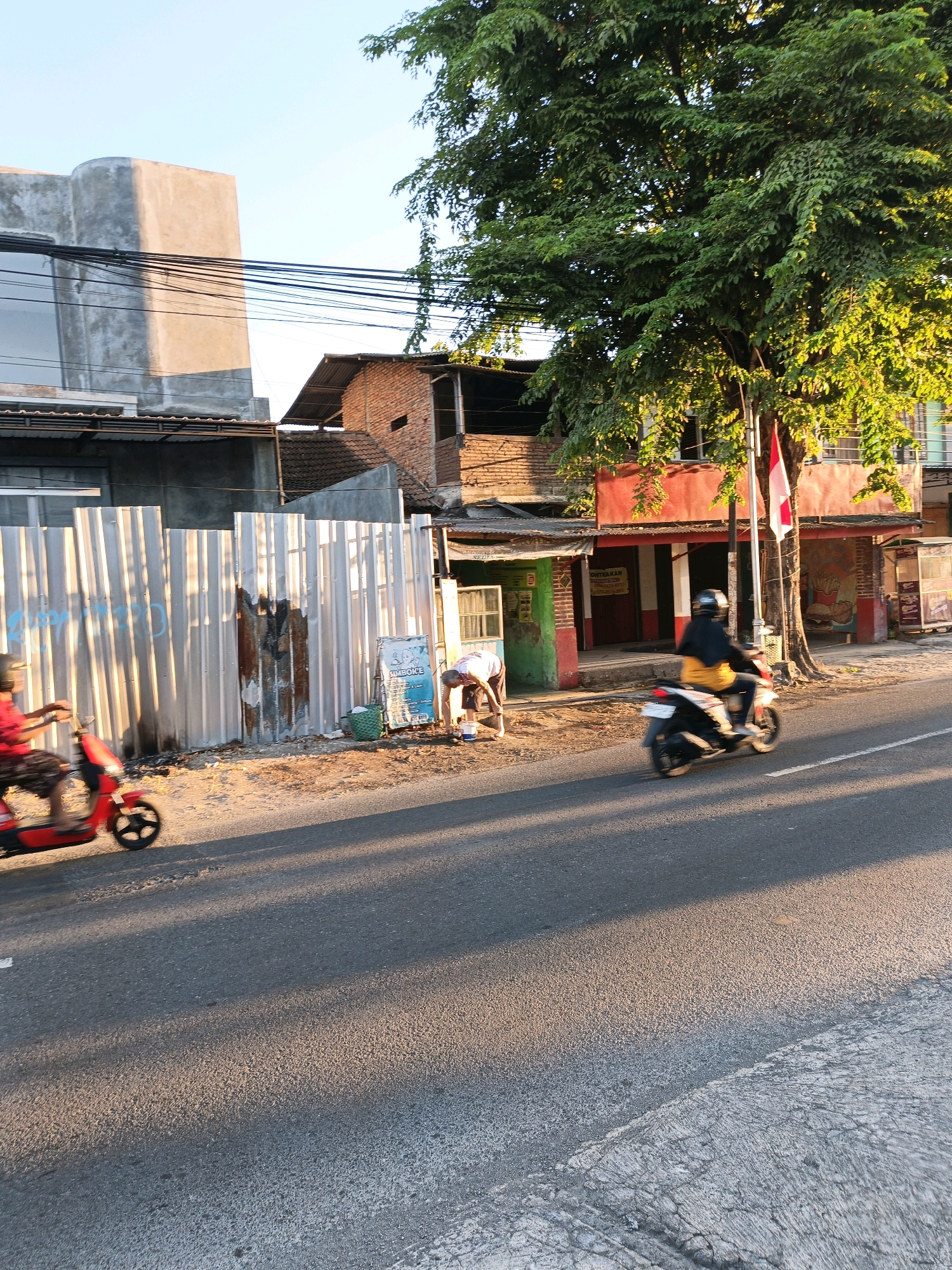 The image depicts a street scene in Klaten, Indonesia, with a focus on a road and buildings. In the foreground, a paved road occupies the majority of the frame, with sunlight casting long shadows across its surface. To the left, a red scooter with a person on it is captured in motion, exhibiting motion blur. Further back on the left, a corrugated metal fence runs along the side of the road, with some weathered sections and graffiti visible. Behind the fence, there are buildings with varying architectural styles, including a modern concrete structure and older brick buildings with corrugated roofs.
On the right side of the image, another person on a scooter, wearing a black helmet and a yellow jacket, is also moving along the road. Adjacent to the road on the right, there is a building with a red facade and an awning. A large tree with lush green foliage stands prominently near the building, its branches extending over the scene. A flag, appearing to be the Indonesian flag, is visible on a flagpole in front of the tree. A person is seen bending down near a blue sign and a green basket, possibly engaged in an activity by the roadside. The overall impression is a snapshot of daily life on a street in Klaten, likely during the late afternoon given the quality of light and shadows.