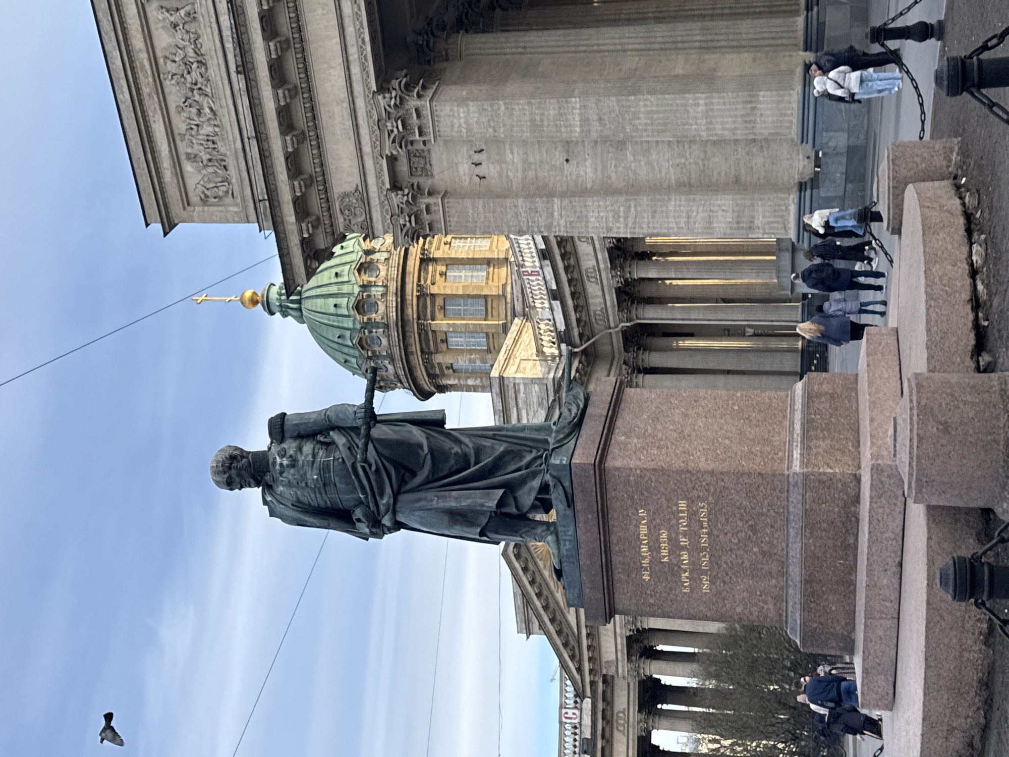 The image shows a statue of Field Marshal Michael Andreas Barclay de Tolly, with the Kazan Cathedral in the background, a major landmark in Saint Petersburg, Russia. The statue stands on a stone plinth, and its inscription is visible. Several people are walking and standing near the cathedral. The sky is partly cloudy. A bird is flying in the lower left of the image. The architecture of the cathedral is prominent, particularly its dome and columns.